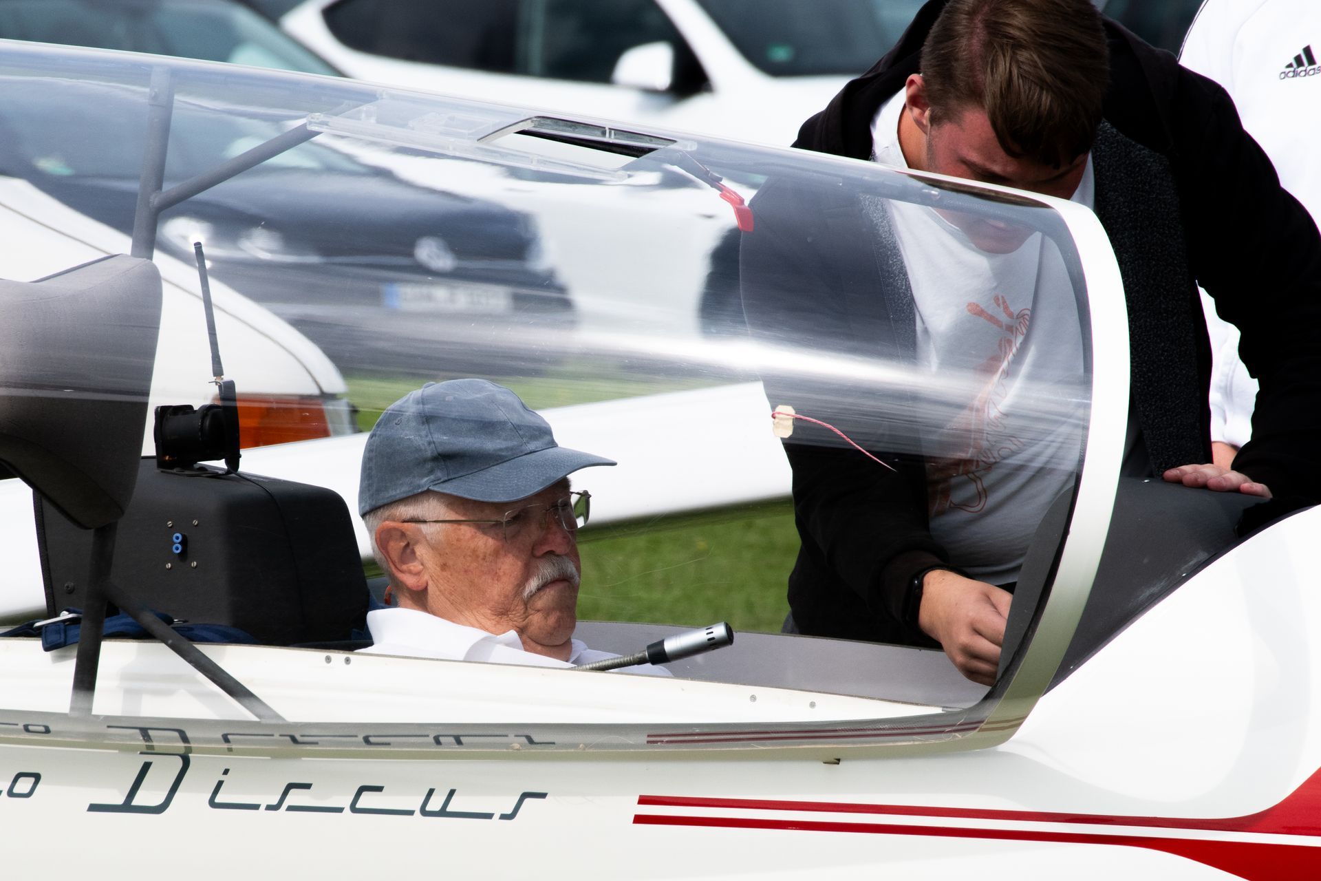 Ein Mann im Cockpit eines Segelflugzeugs wird von einer anderen Person unterstützt. Die Szenerie scheint im Freien zu spielen, möglicherweise auf einem Flugfeld.