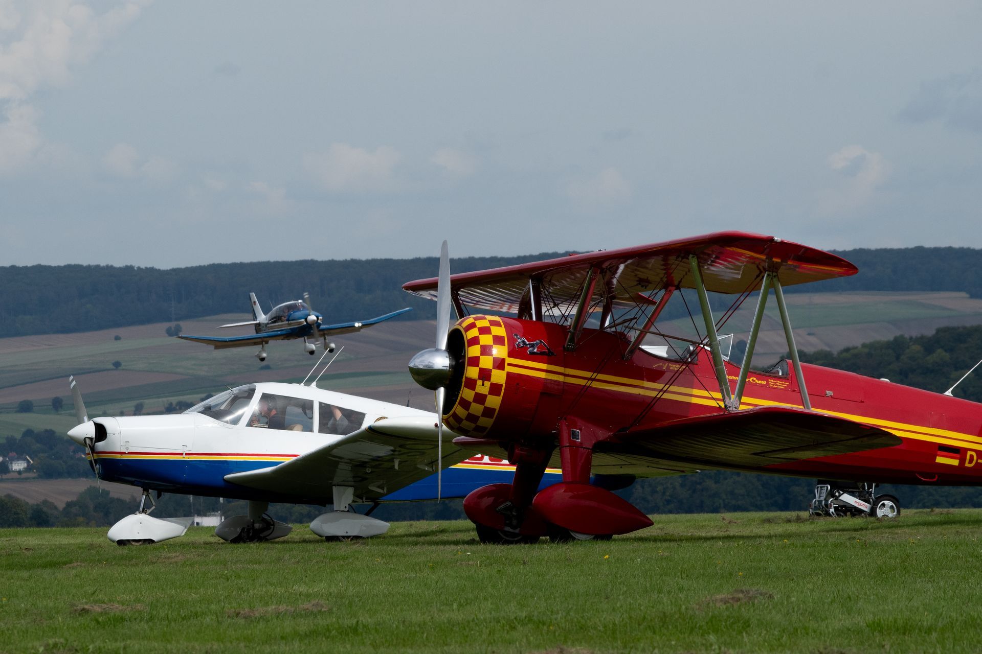 Drei Flugzeuge auf einem grasbewachsenen Feld; ein rotes Doppeldeckerflugzeug vorne, daneben ein weiß-blaues Flugzeug, im Hintergrund ein startendes Flugzeug.