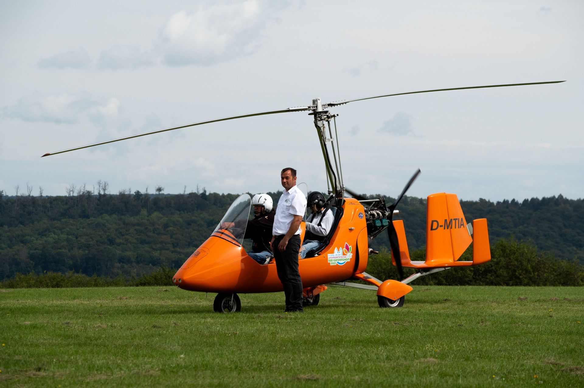Ein Mann steht neben einem orangefarbenen Tragschrauber auf einem grasbewachsenen Feld; im Hintergrund sind Bäume zu sehen.