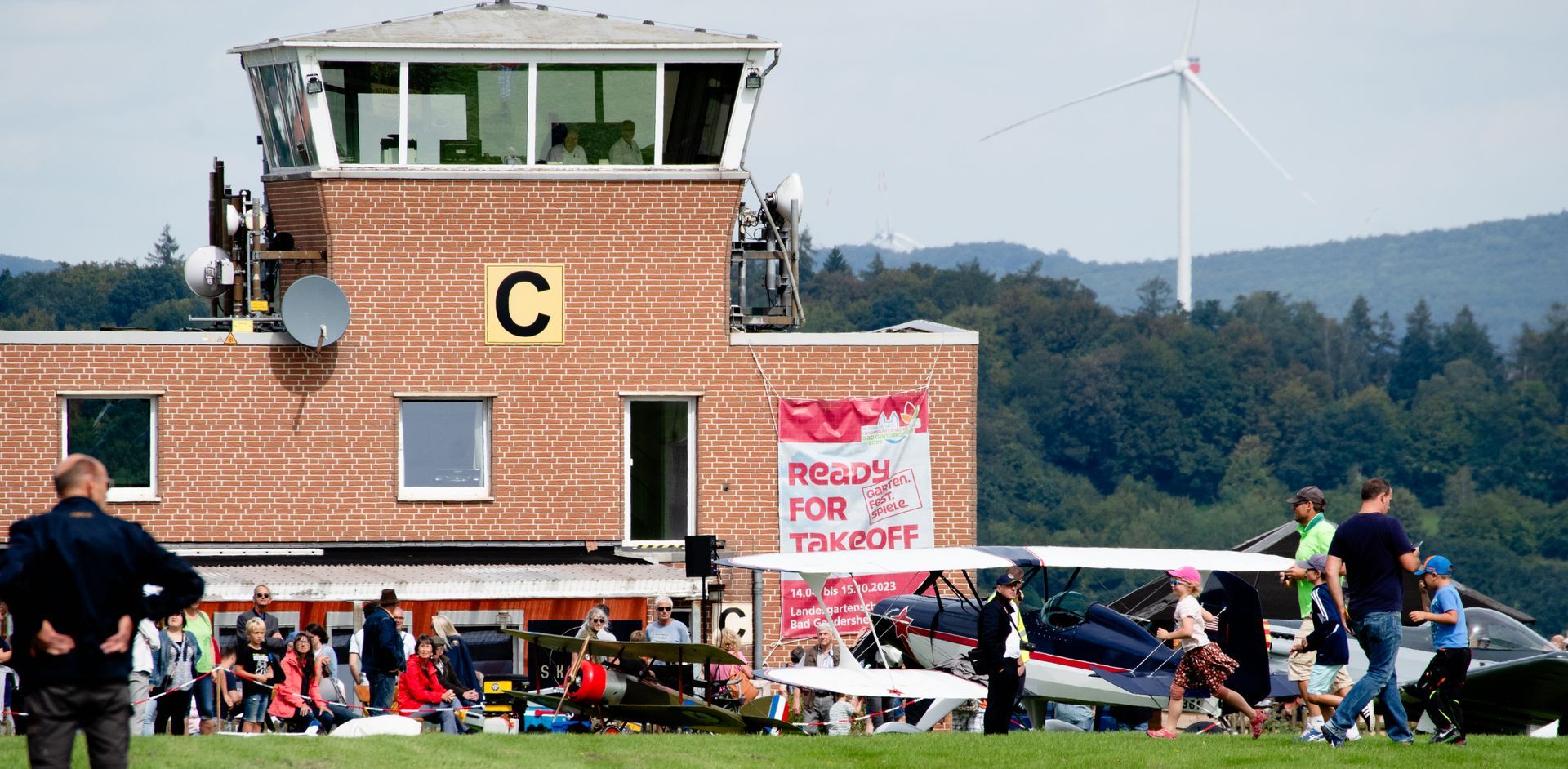 Ein Flughafen-Kontrollturm mit einer Menschenmenge auf einem Rasenfeld; im Hintergrund ist eine Windkraftanlage zu sehen.