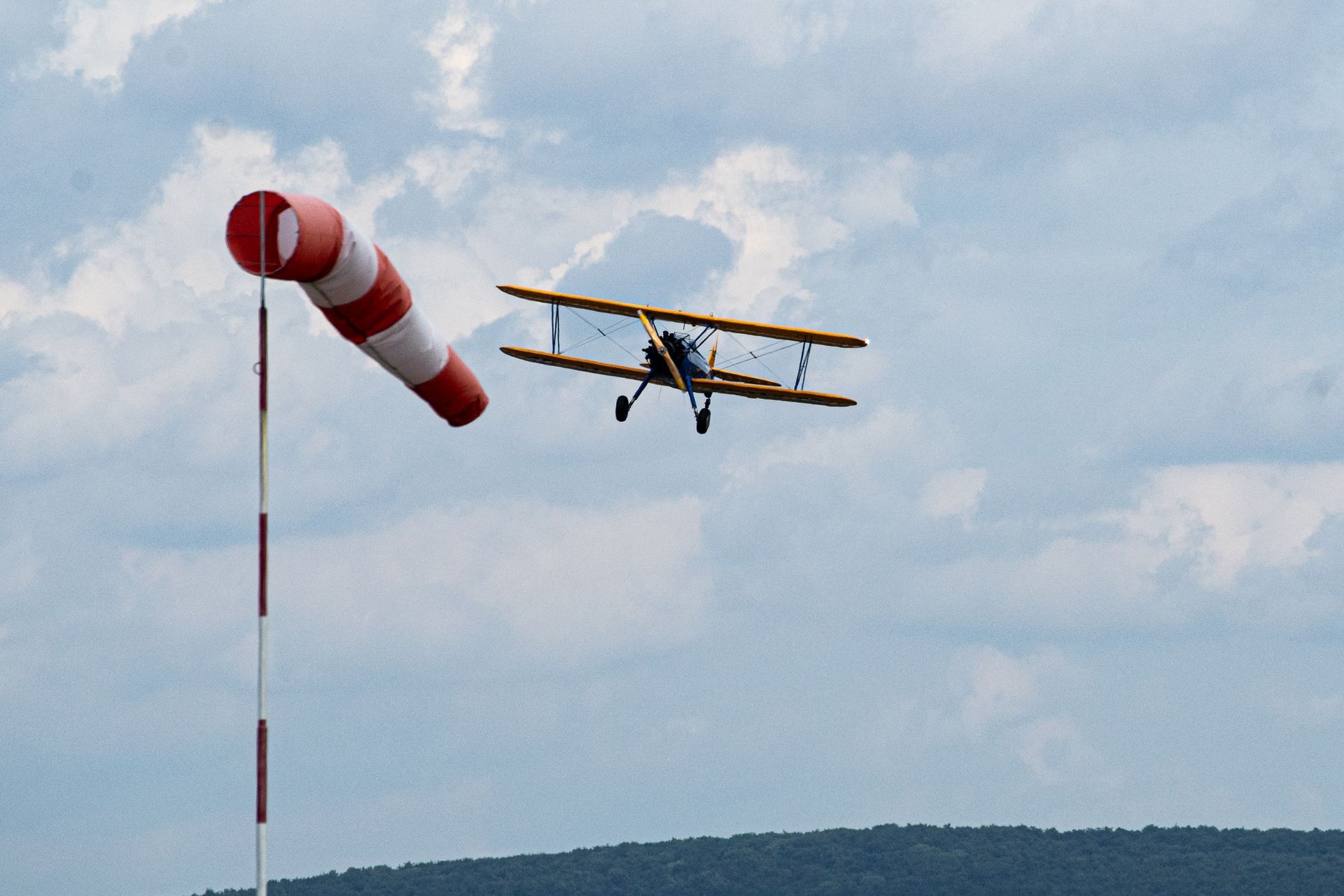 Doppeldecker fliegt in der Nähe eines Windsacks; rot-weiße Streifen auf dem Windsack, gelb-blaues Flugzeug, bewölkter Himmel.