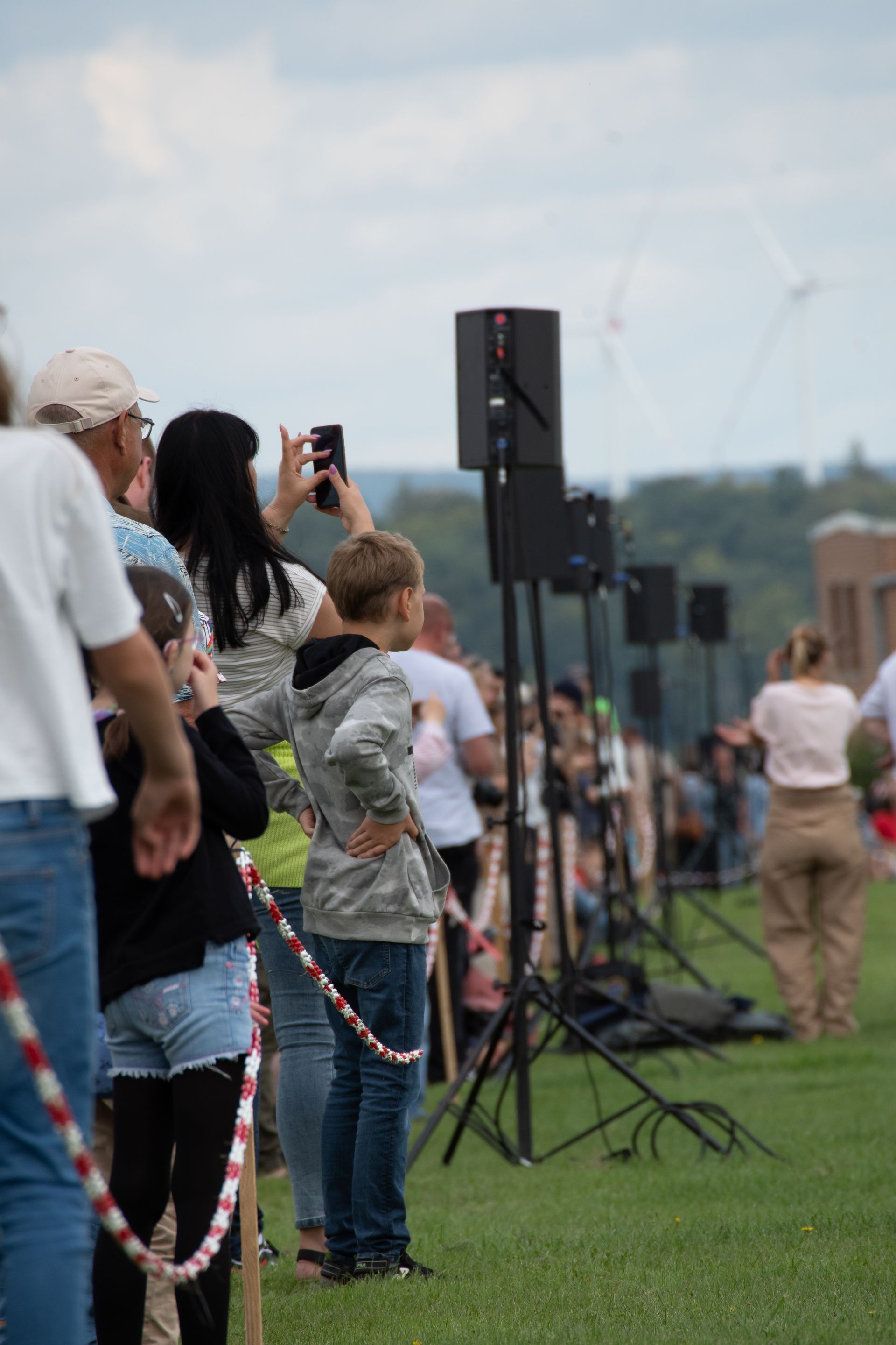 Zuschauer beobachten die Veranstaltung; Menschenmenge hinter Absperrseil, Redner auf Stativen. Grünes Gras, bewölkter Himmel, einige machen Fotos.