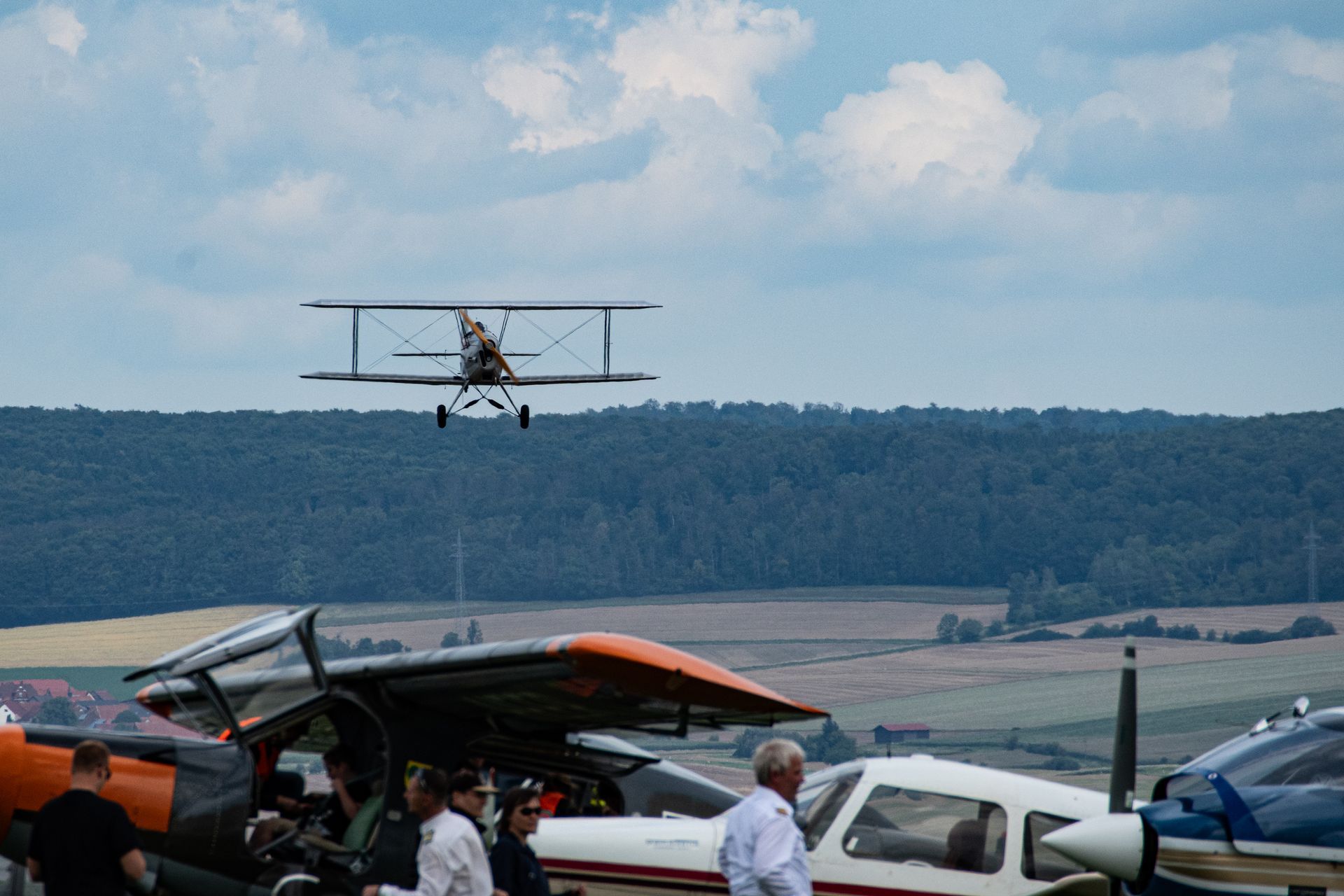 Ein Doppeldecker startet über geparkten Flugzeugen und einer Gruppe von Menschen auf einem Flugfeld.