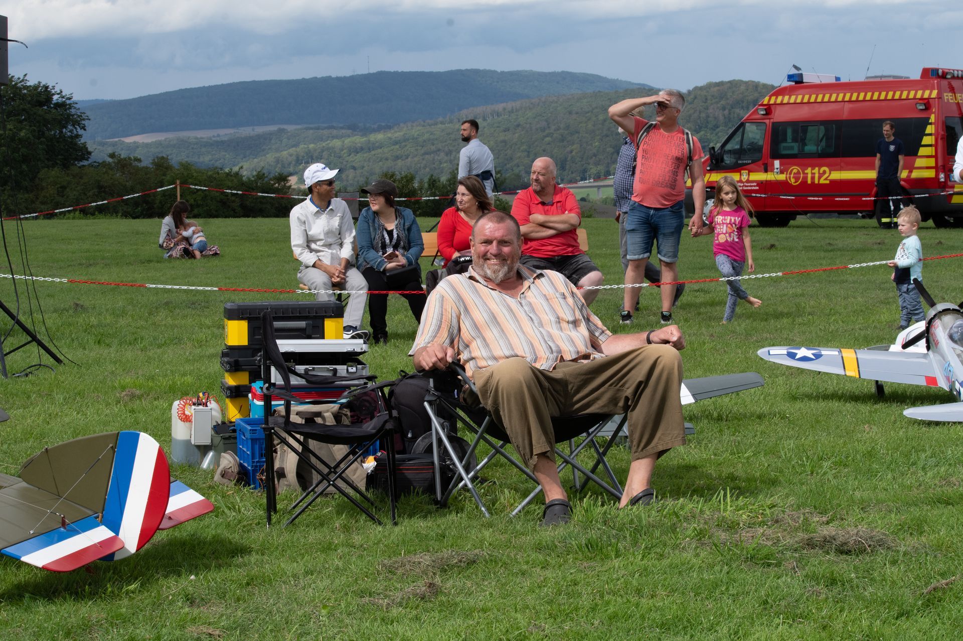 Ein Mann auf einem Klappstuhl beobachtet eine Veranstaltung mit ferngesteuerten Flugzeugen auf einer Wiese, während Zuschauer und ein Einsatzfahrzeug anwesend sind.