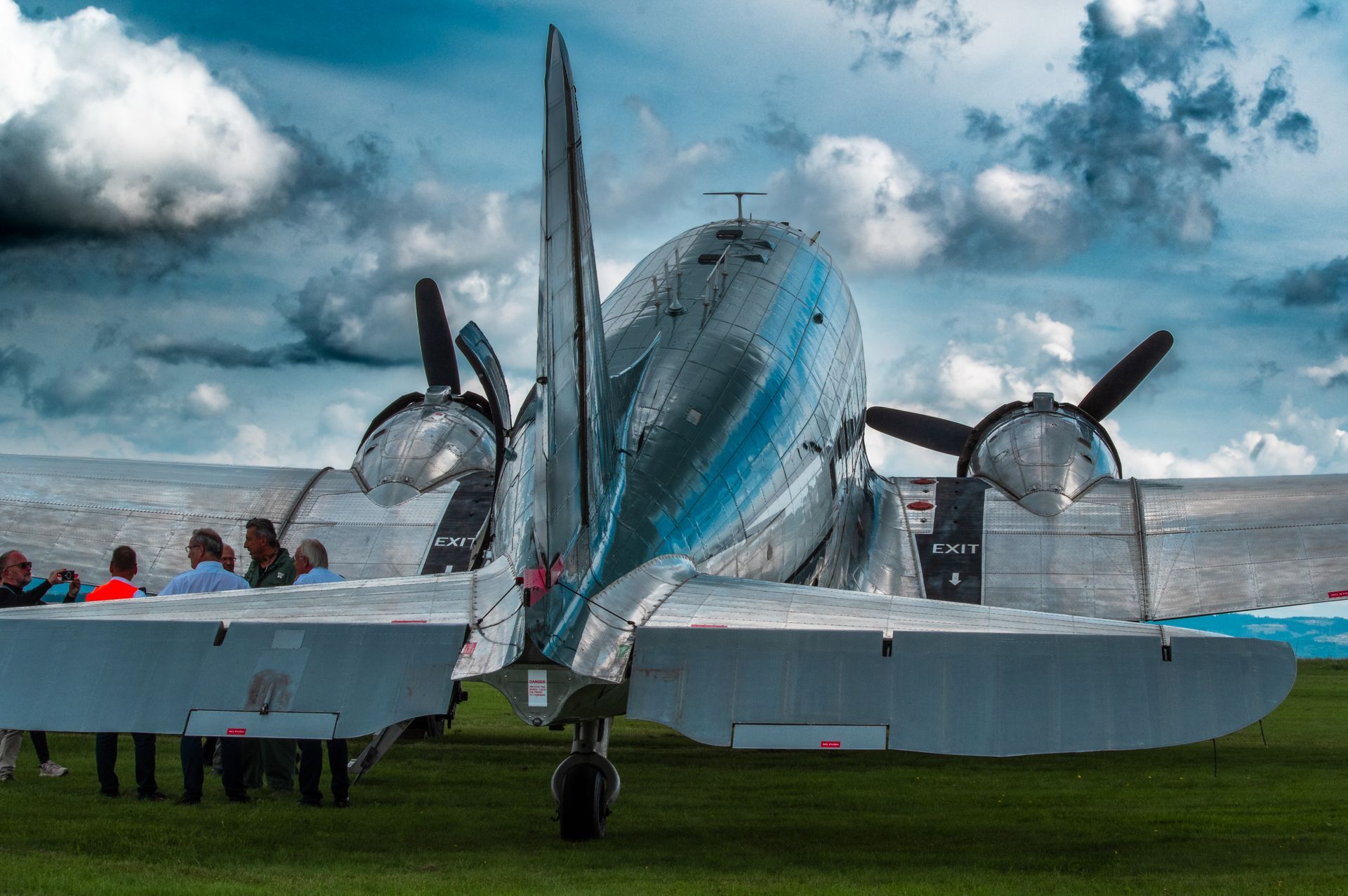 Rückansicht eines glänzenden Oldtimer-Flugzeugs auf einem grasbewachsenen Feld mit Menschen im Hintergrund unter bewölktem Himmel.