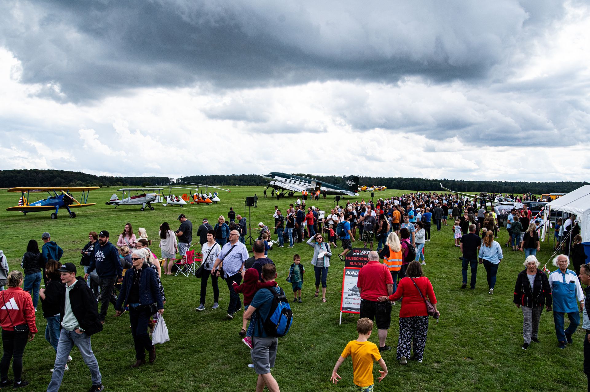 Menschenmenge bei einer Flugschau auf einem grasbewachsenen Flugfeld; Flugzeuge geparkt, bedeckter Himmel.