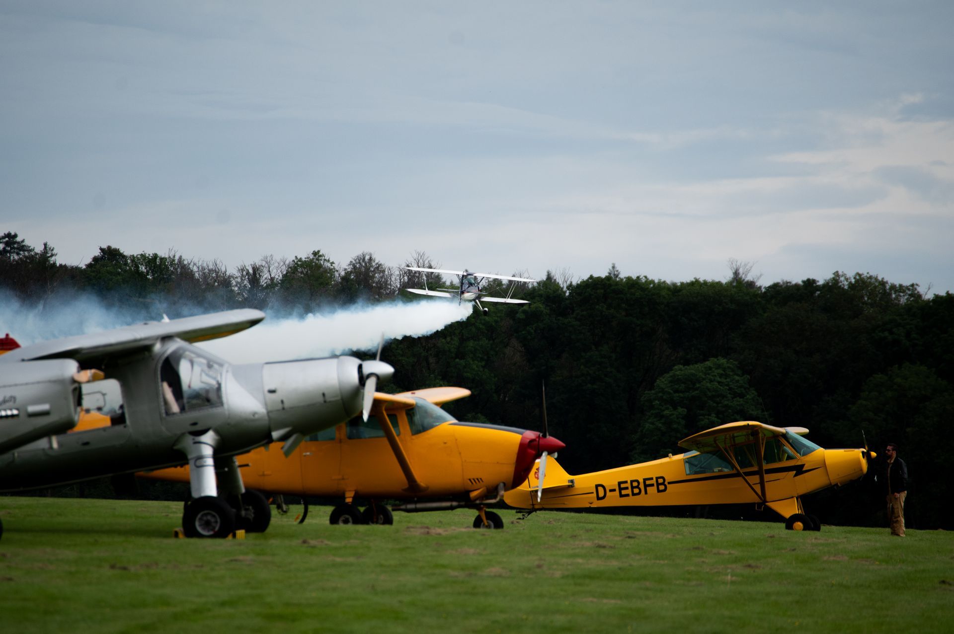 Drei Flugzeuge auf einem Grasfeld; eines davon mit Rauch. Bäume im Hintergrund, bewölkter Himmel.
