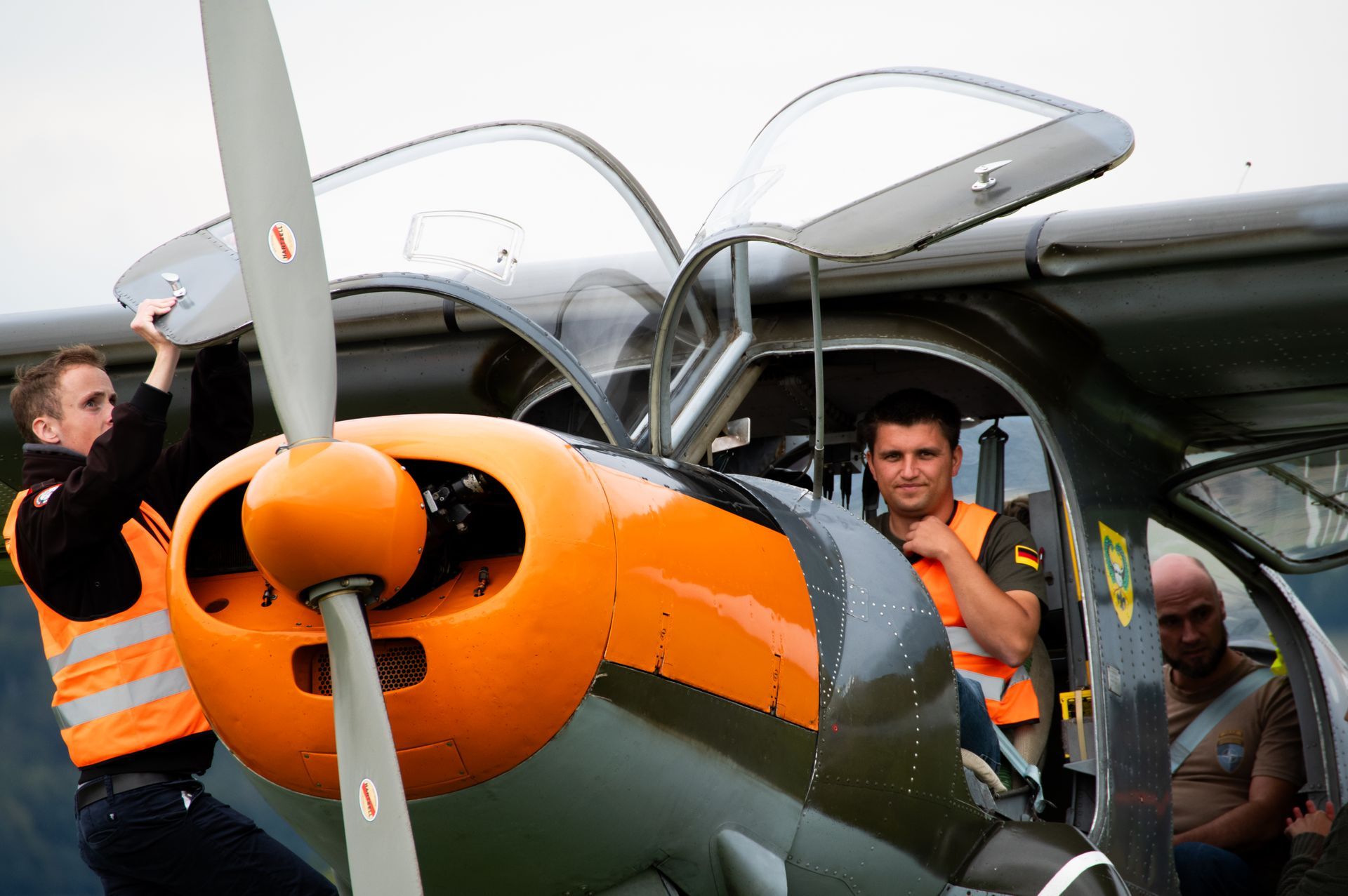 Drei Männer in der Nähe eines Propellerflugzeugs. Einer öffnet das Cockpit, zwei sitzen im Cockpit und tragen Sicherheitswesten; das Flugzeug ist orange und grau.
