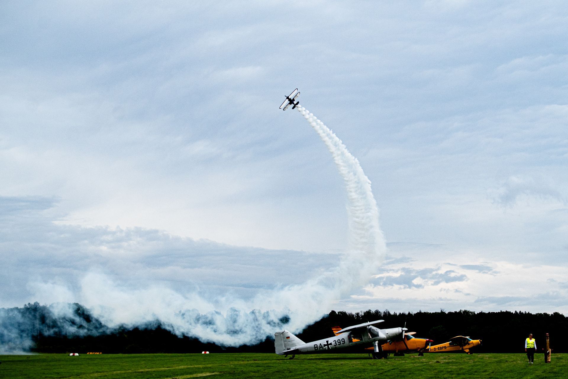 Ein Flugzeug führt einen Kunstflug durch und hinterlässt eine Rauchspur. Mehrere Flugzeuge sind auf einem Grasfeld geparkt.