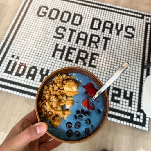 Una mano sostiene un bol azul de smoothie cubierto con granola, arándanos y fresas sobre una esterilla