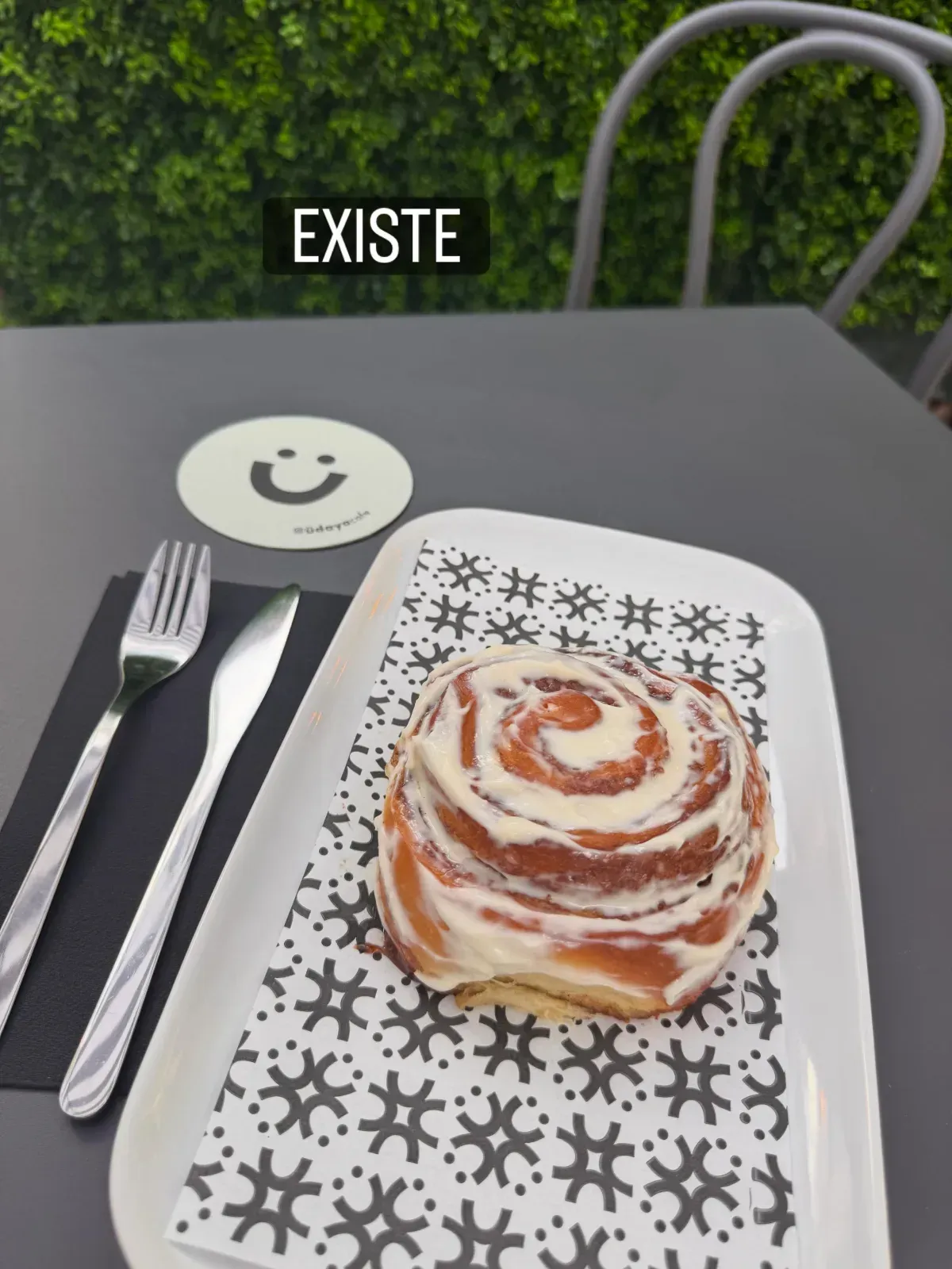 Un rollo de canela en un plato estampado, con tenedor y cuchillo, sobre una mesa negra al aire libre;