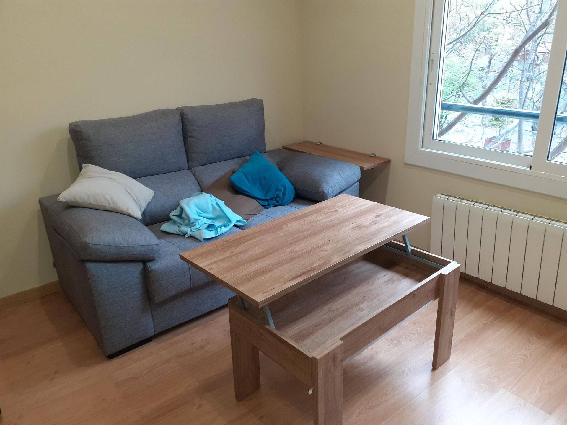 Una sala de estar con un sofá gris, una mesa de centro elevable de madera y una ventana con radiador.