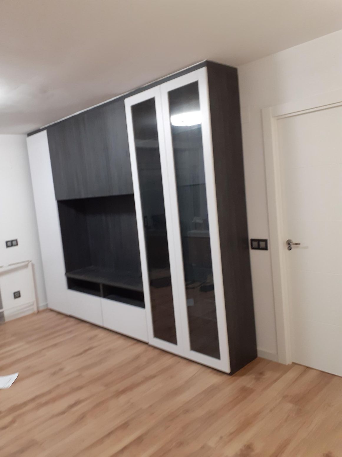 Mueble empotrado con paneles en gris oscuro y blanco, puertas de cristal y nicho para TV en estancia con suelo de madera.