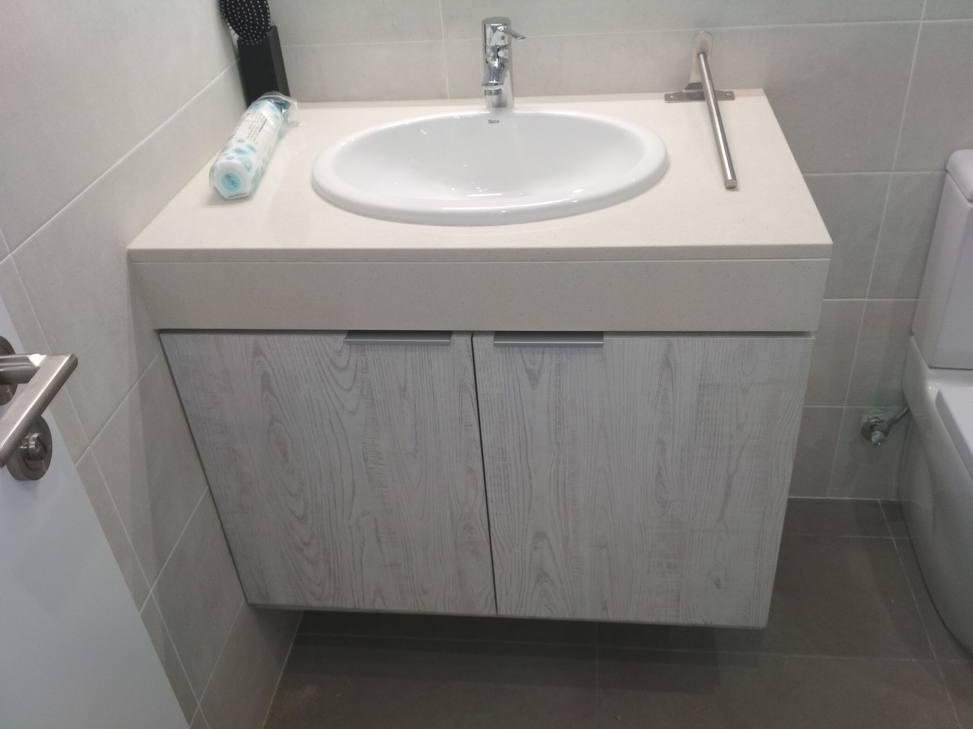 Tocador de baño con lavabo ovalado, puertas de gabinete con veta de madera clara y herrajes cromados.