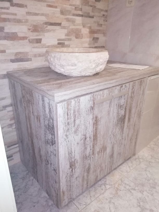 Tocador de baño con lavabo de piedra sobre un mueble de madera envejecida, contra una pared de azulejos de piedra.