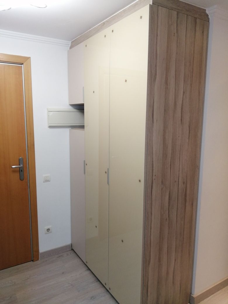 Armario empotrado con paneles de madera, puertas beige y una estantería abierta. Paredes claras y puerta de madera.