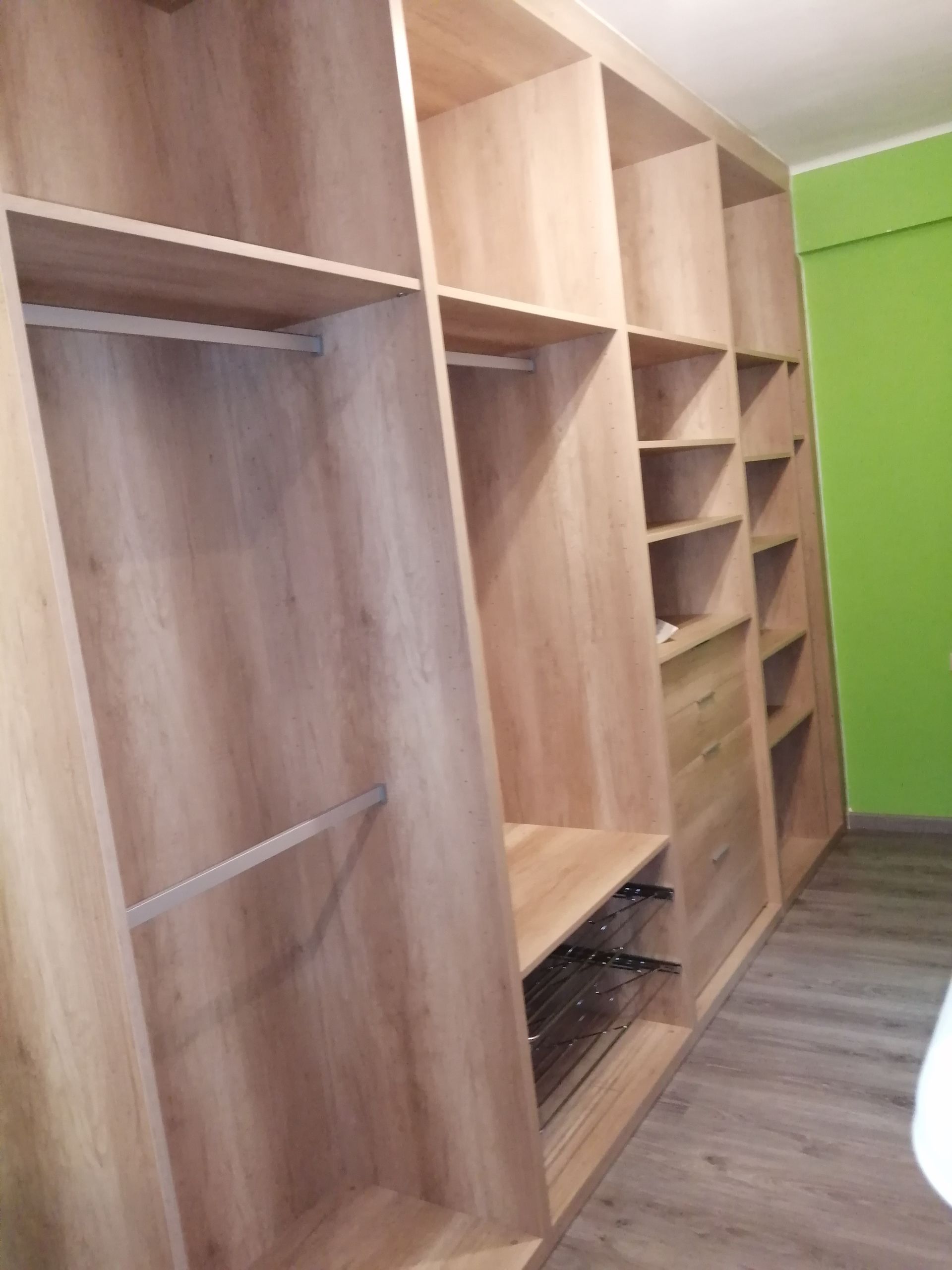 Armario empotrado de madera con estantes, cajones y barras para colgar; colocado contra una pared verde y un piso de madera.