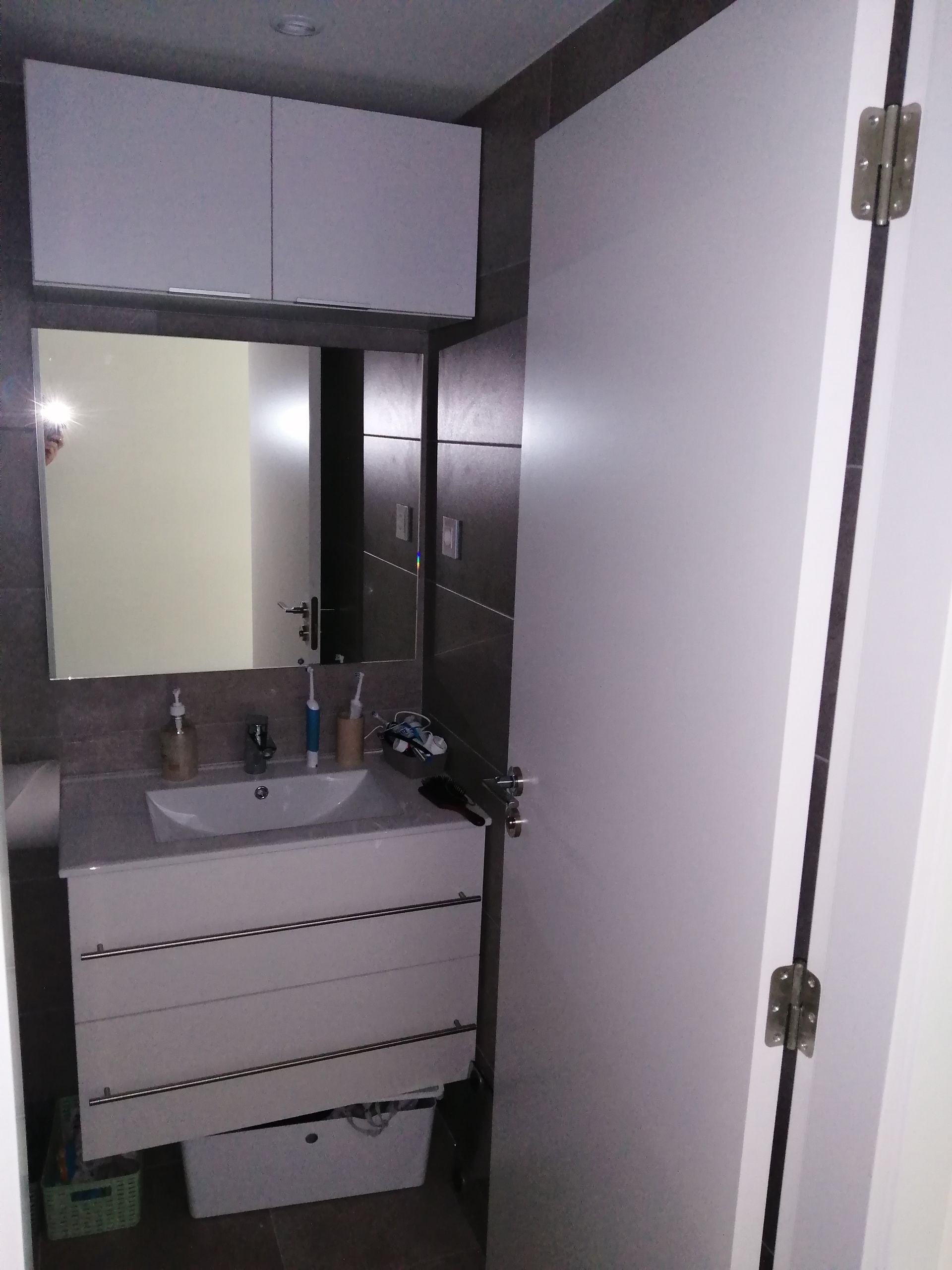 Pequeño baño blanco con lavabo, mueble, espejo y puerta blanca cerrada.