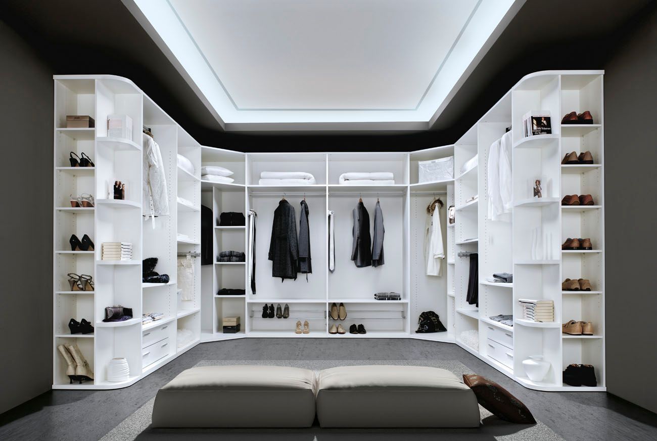 Vestidor blanco con ropa, zapatos y un banco. Paredes grises, techo blanco con iluminación empotrada.