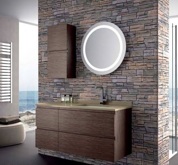 Baño con pared con acento de piedra, tocador marrón, espejo redondo iluminado y vista al mar.
