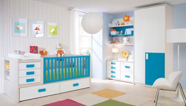 Una habitación infantil luminosa con cuna, cómoda y armario en blanco y azul. Obras de arte coloridas y una alfombra estampada decoran la habitación.