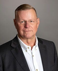 Ein Mann in Anzug und weißem Hemd steht vor grauem Hintergrund.