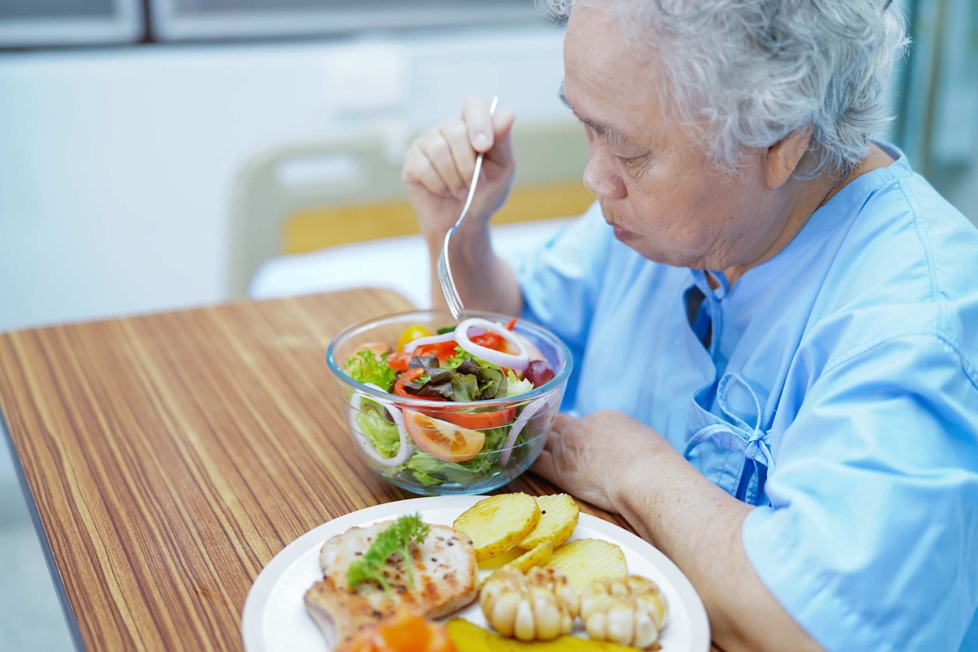 Persona mayor comiendo ensalada y comida en una habitación de hospital, vestida con una bata azul.