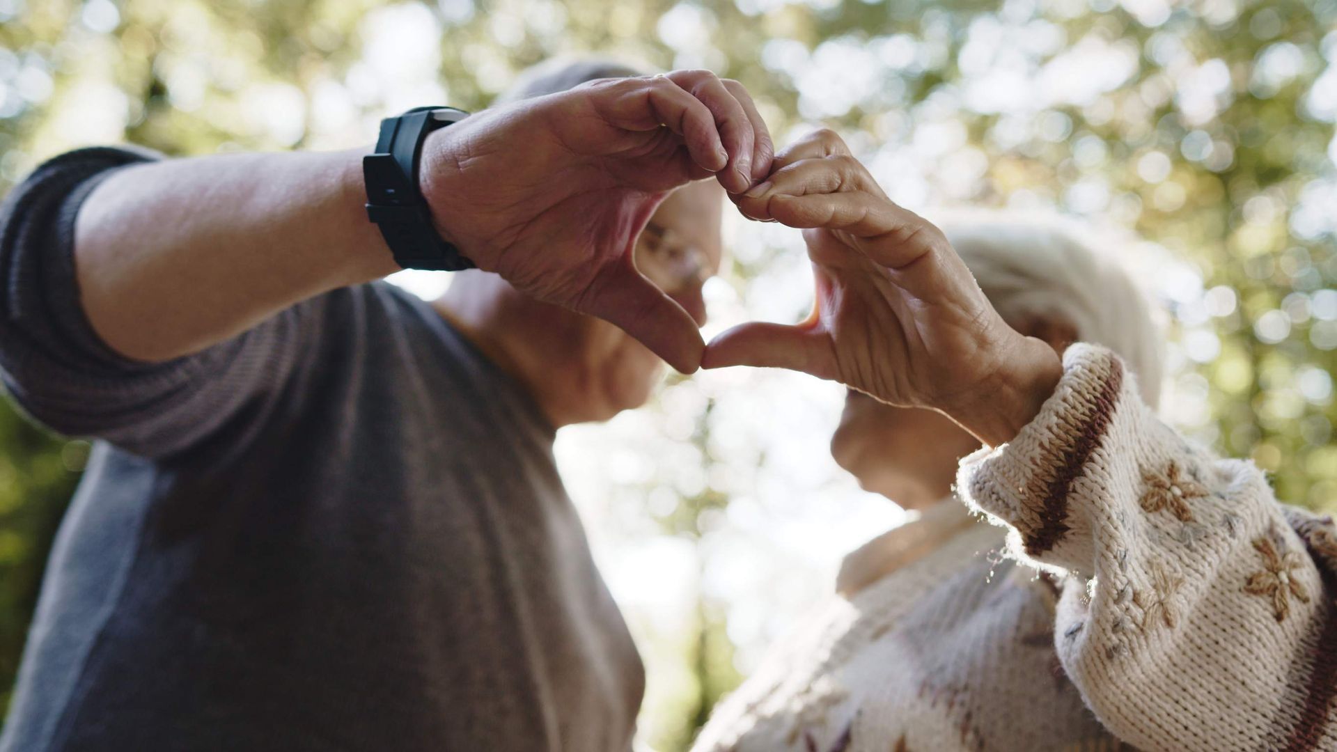 Una pareja de ancianos formando un corazón con sus manos en un entorno al aire libre.