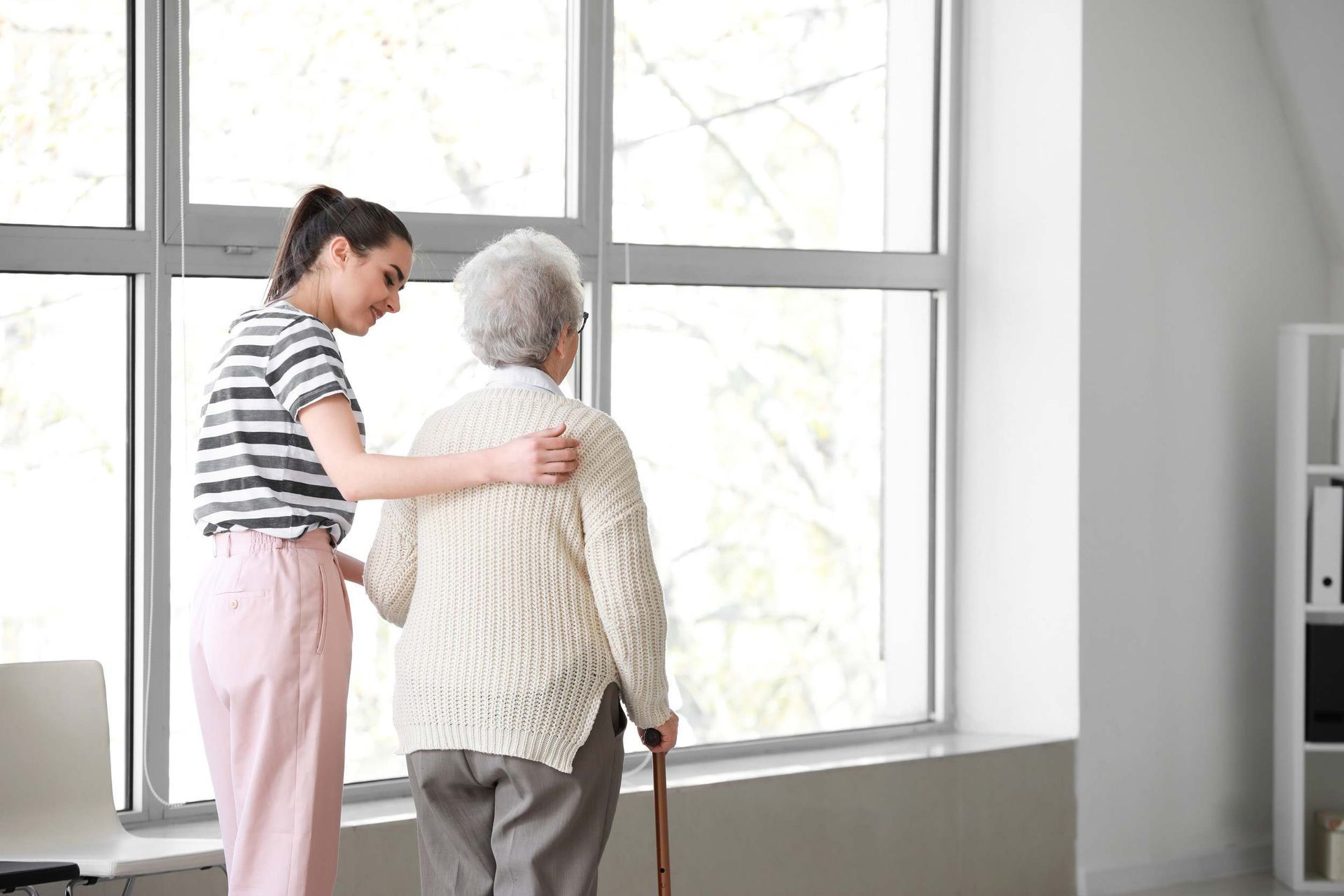 Una mujer joven ayuda a una anciana con un bastón a mirar por la ventana.