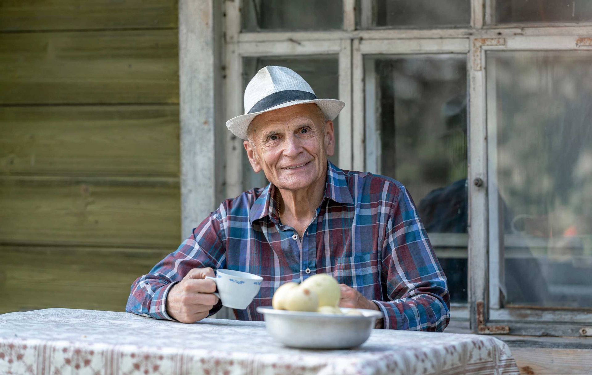 Un hombre mayor, con camisa a cuadros y sombrero, sonríe mientras sostiene una taza, sentado en una mesa con fruta.