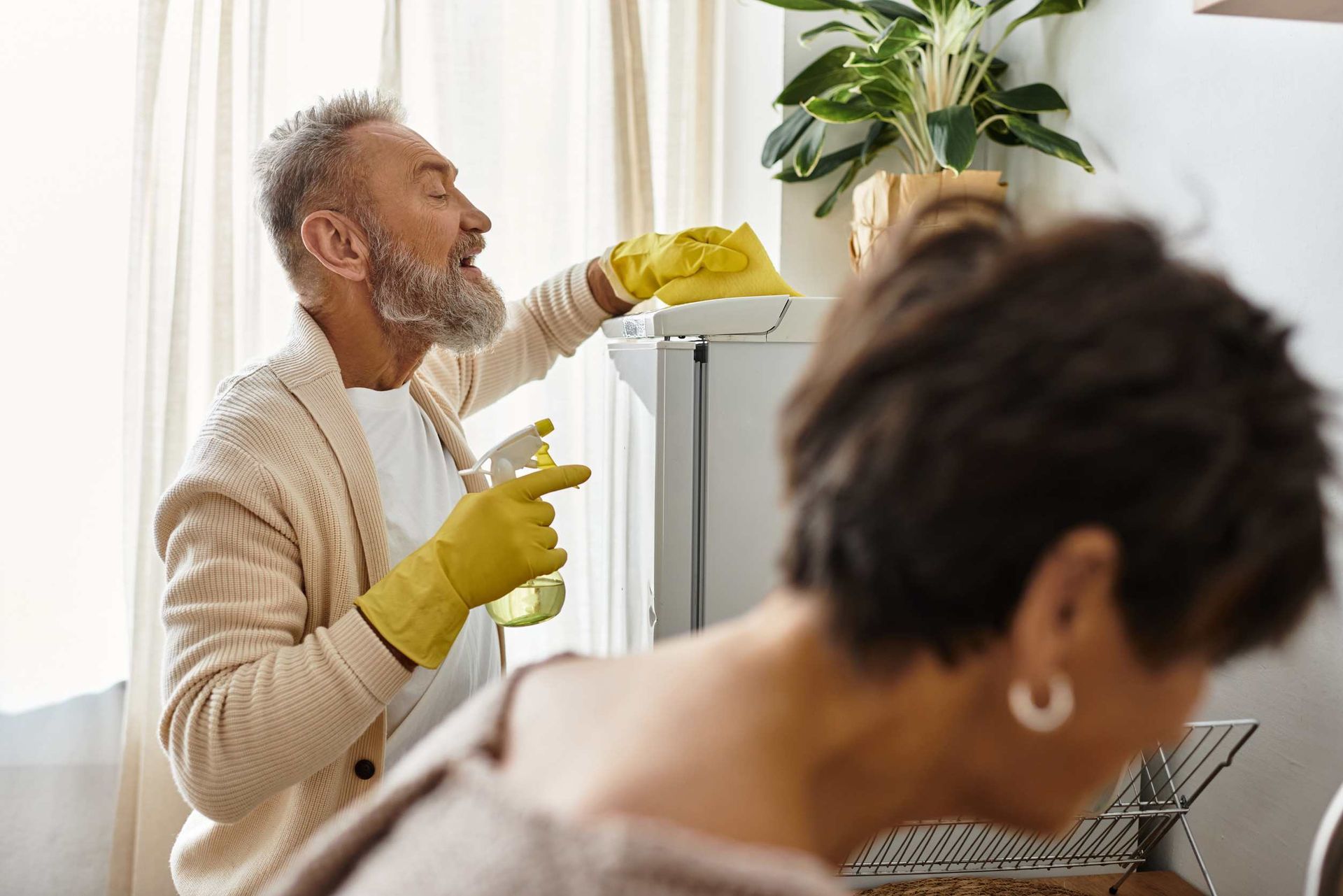 Hombre con guantes limpiando un refrigerador; mujer en primer plano, planta en el fondo.