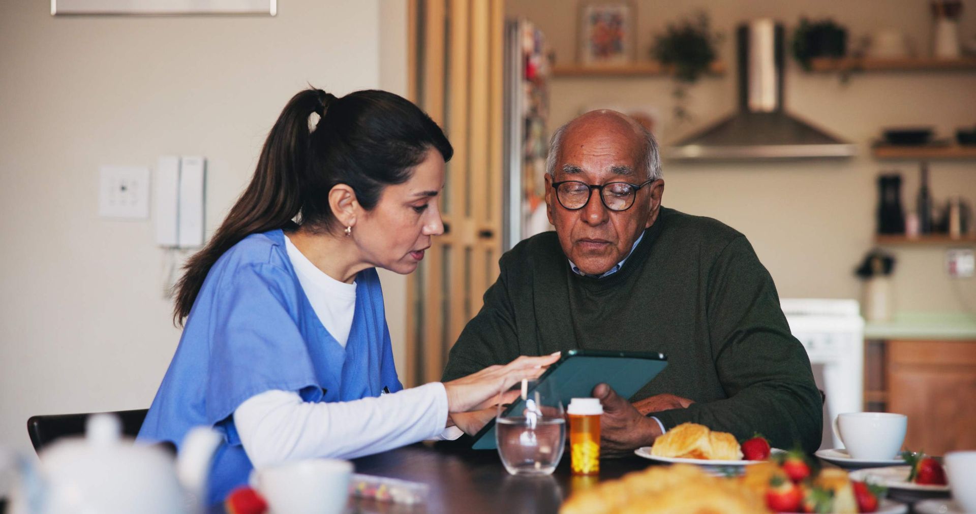 Una cuidadora con uniforme médico señala una tableta y le explica a un anciano sentado a la mesa. Se ve el desayuno.