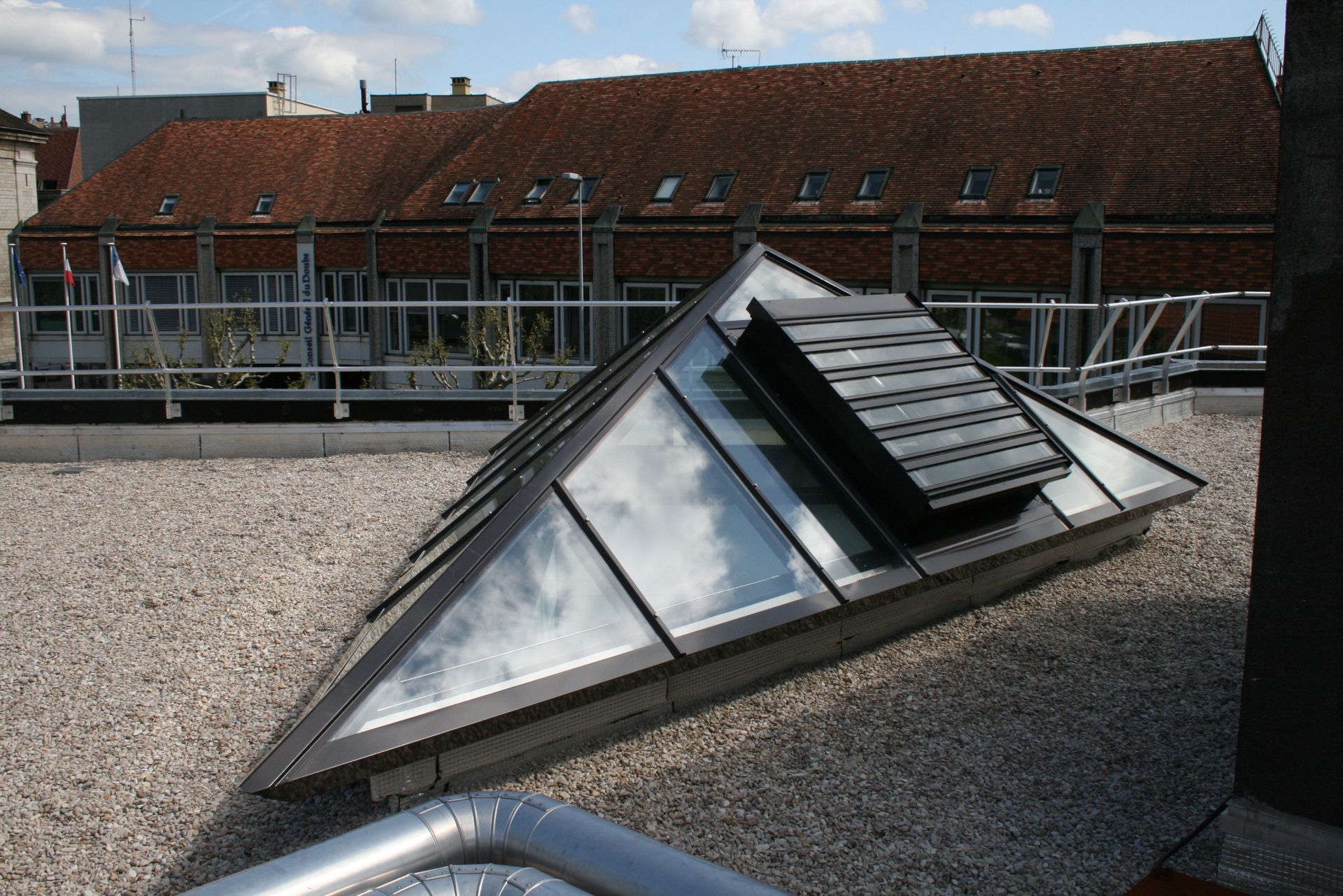 Puits de lumière pyramidal sur un toit de gravier, avec un bâtiment en arrière-plan. Le ciel bleu se reflète dans la vitre.
