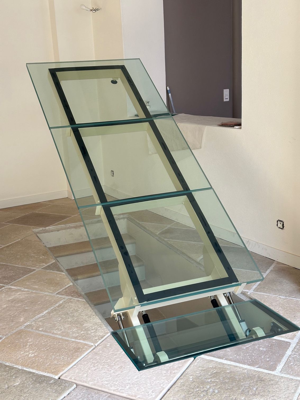 Plancher vitré, articulé et ouvrant sur un espace souterrain. Cadres noirs, sol en pierre beige, décor intérieur.