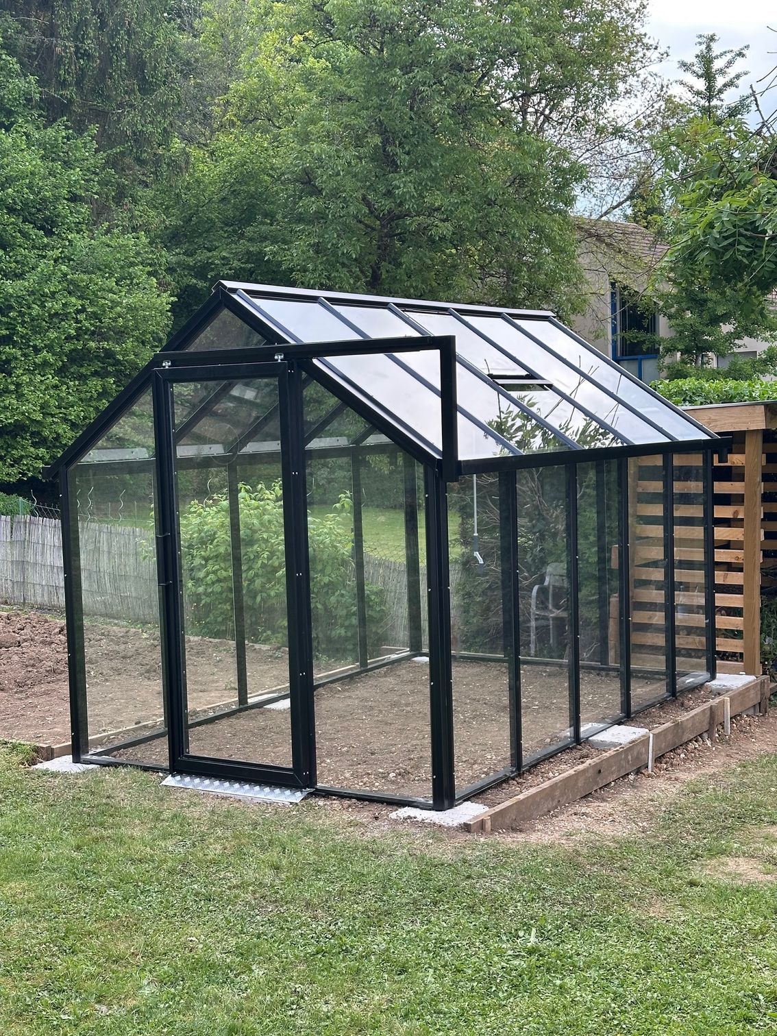 Serre à structure noire et panneaux de verre, installée sur un sol en gravier, dans un jardin.