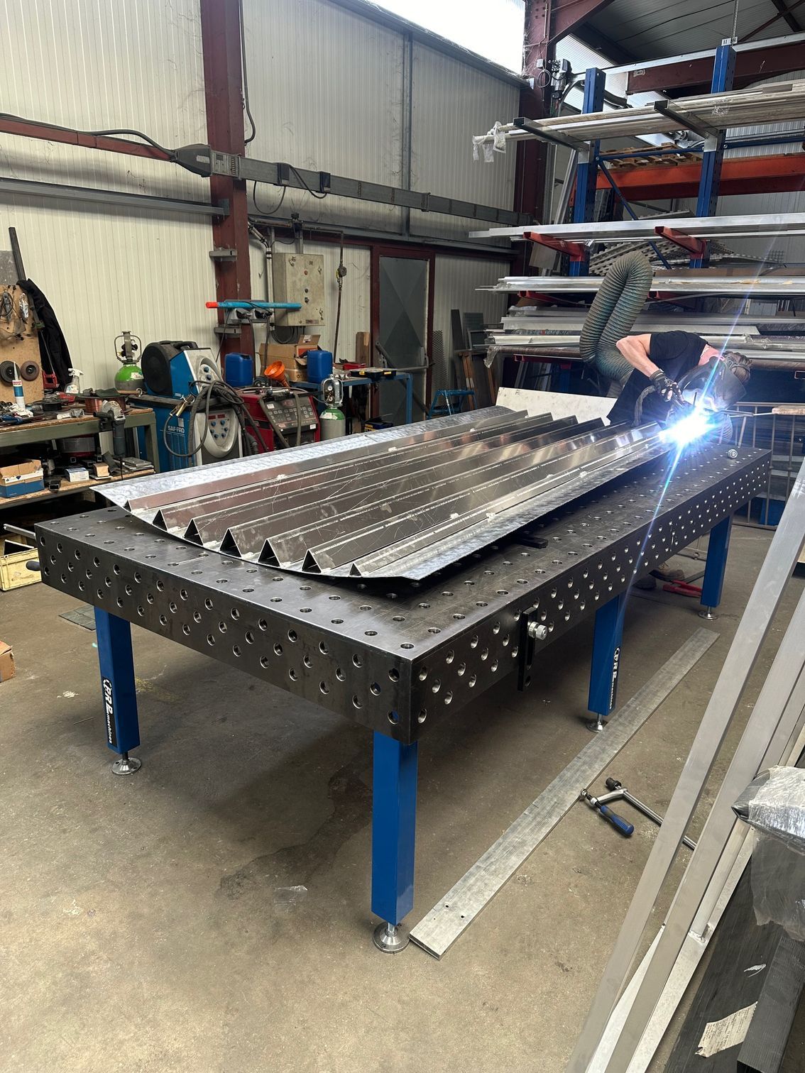 Un soudeur au travail, une torche créant des étincelles sur des tôles métalliques posées sur une grande table de soudage à surface quadrillée à l'intérieur d'un atelier.