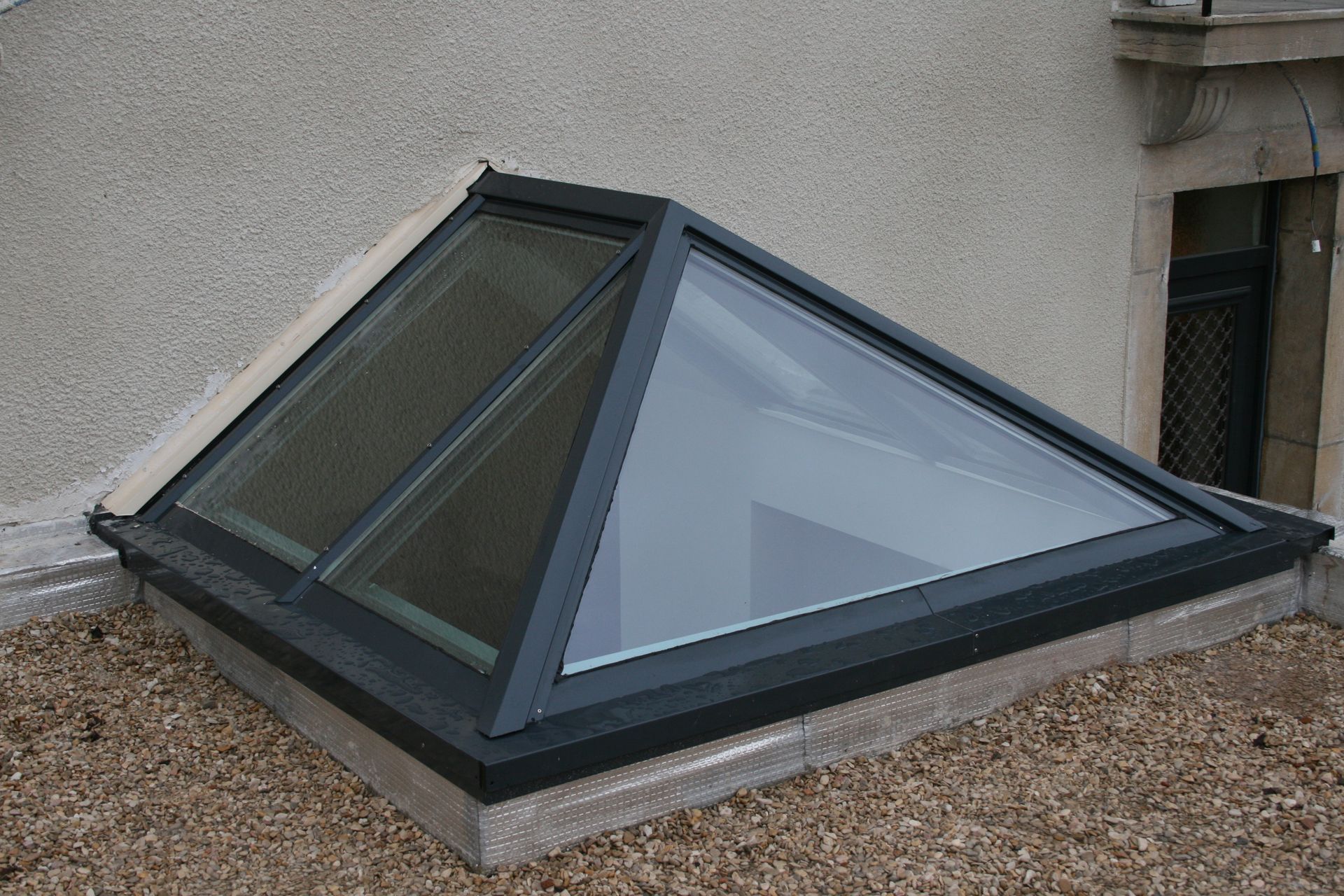 Une lucarne triangulaire à cadre sombre encastrée dans un toit clair, entourée de petits graviers.