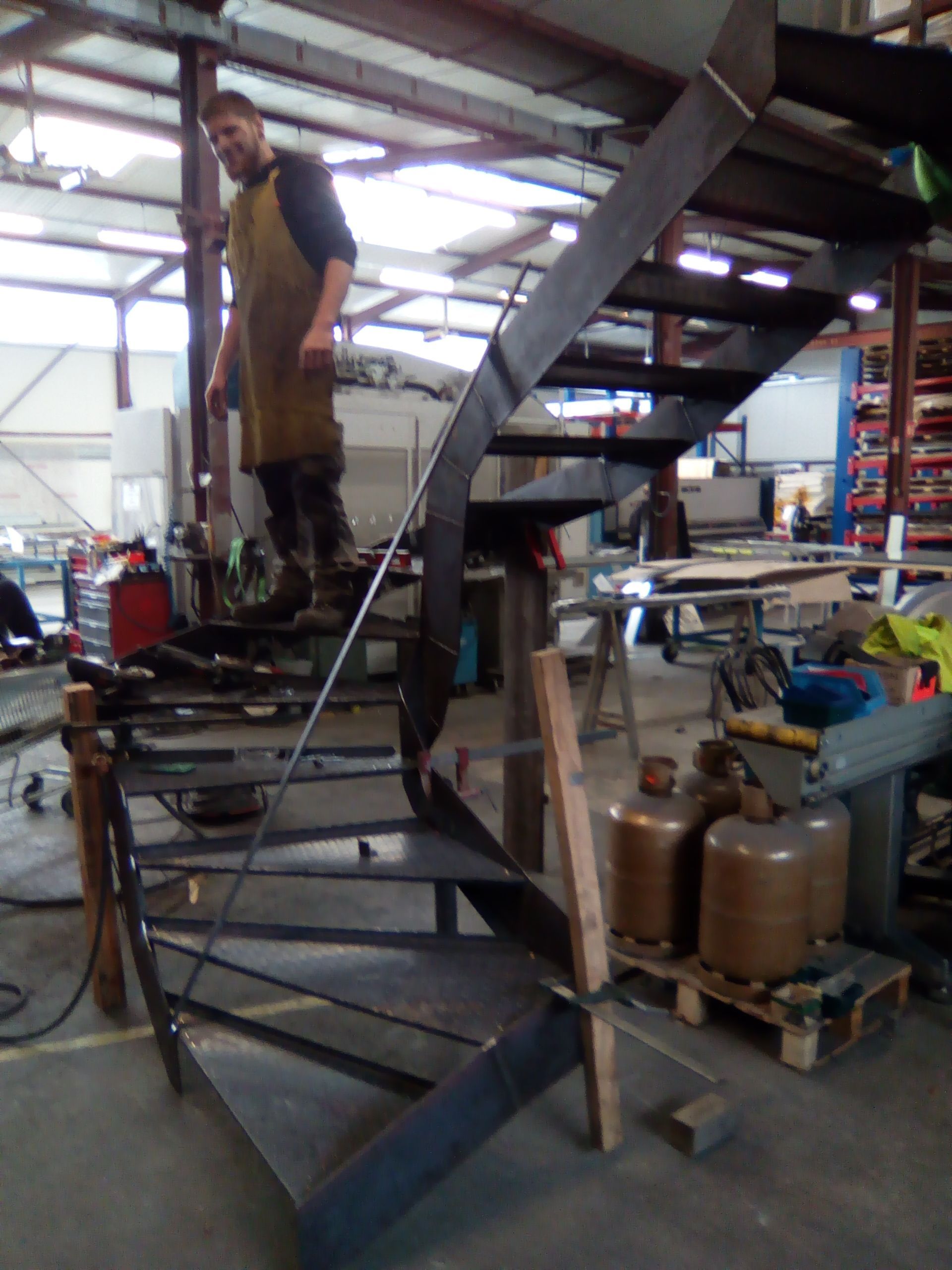 Personne portant un tablier, debout sur un escalier en colimaçon métallique dans un atelier.