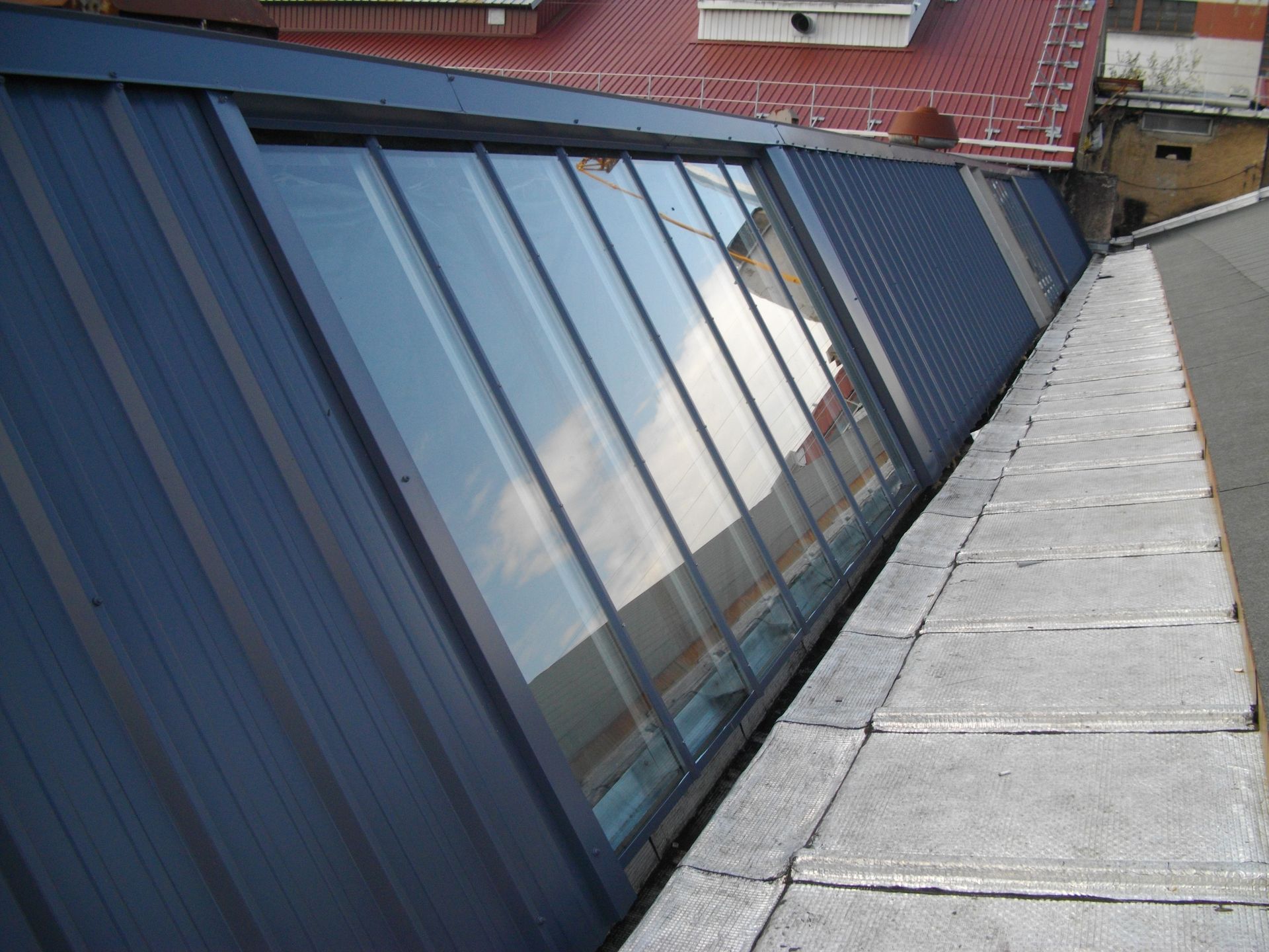 Toit en pente avec une rangée de fenêtres, bardage métallique bleu et corniche en béton.