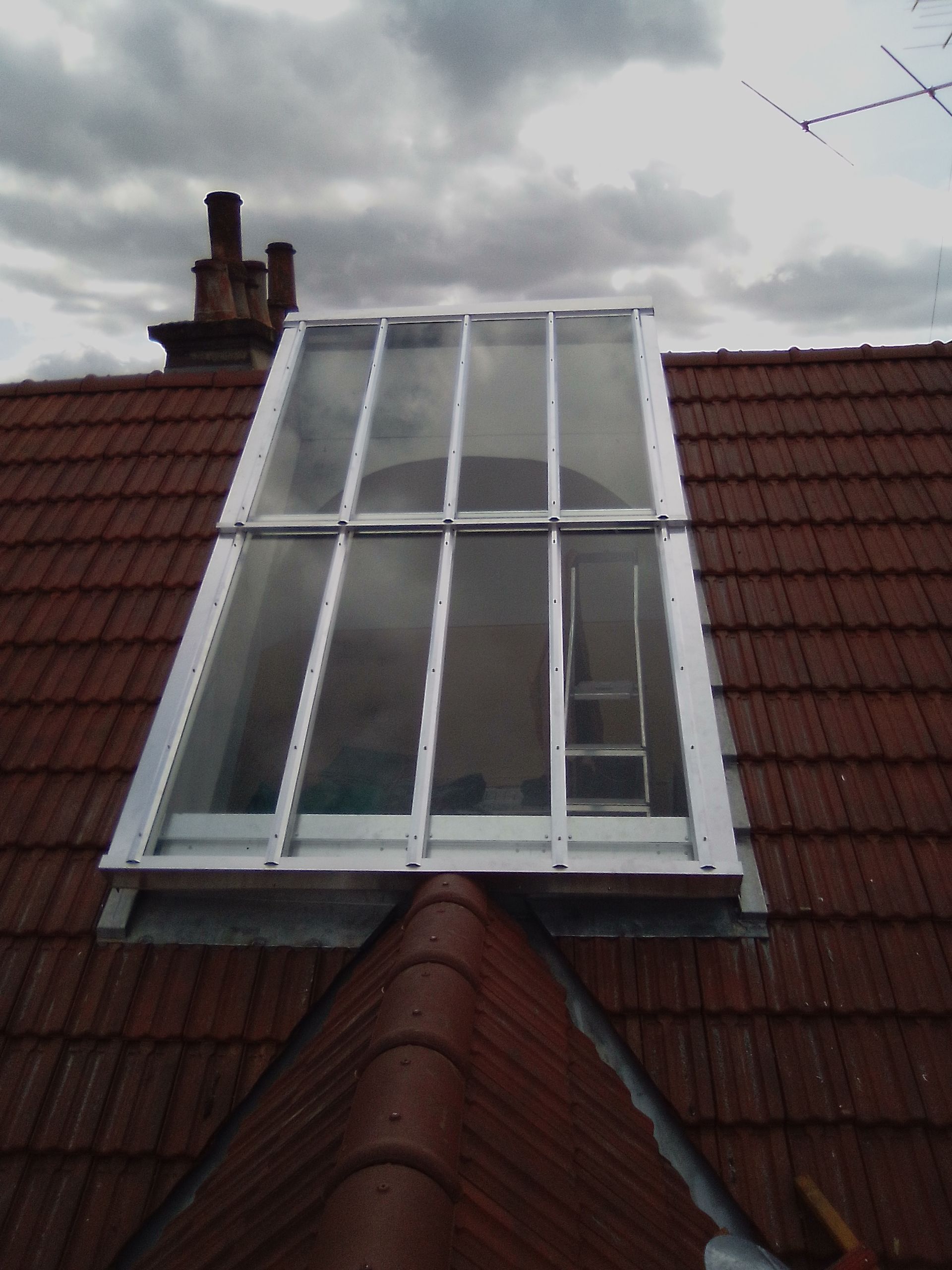 Une lucarne rectangulaire sur un toit de tuiles rouges, avec une cheminée à gauche, sous un ciel nuageux.