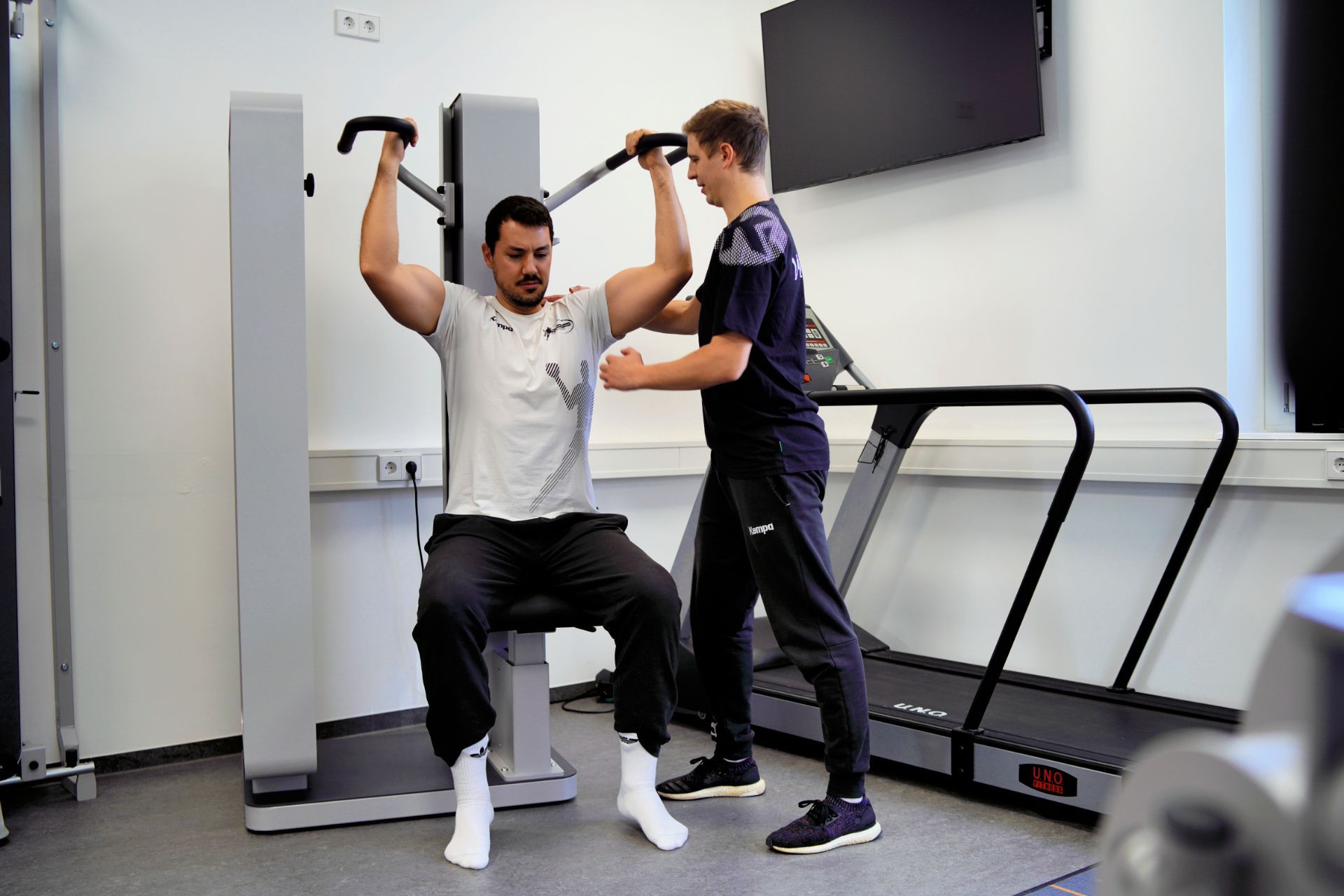 Ein Mann bei einer physiotherapeutischen Übung an einem Gerät unter Aufsicht eines Physiotherapeuten