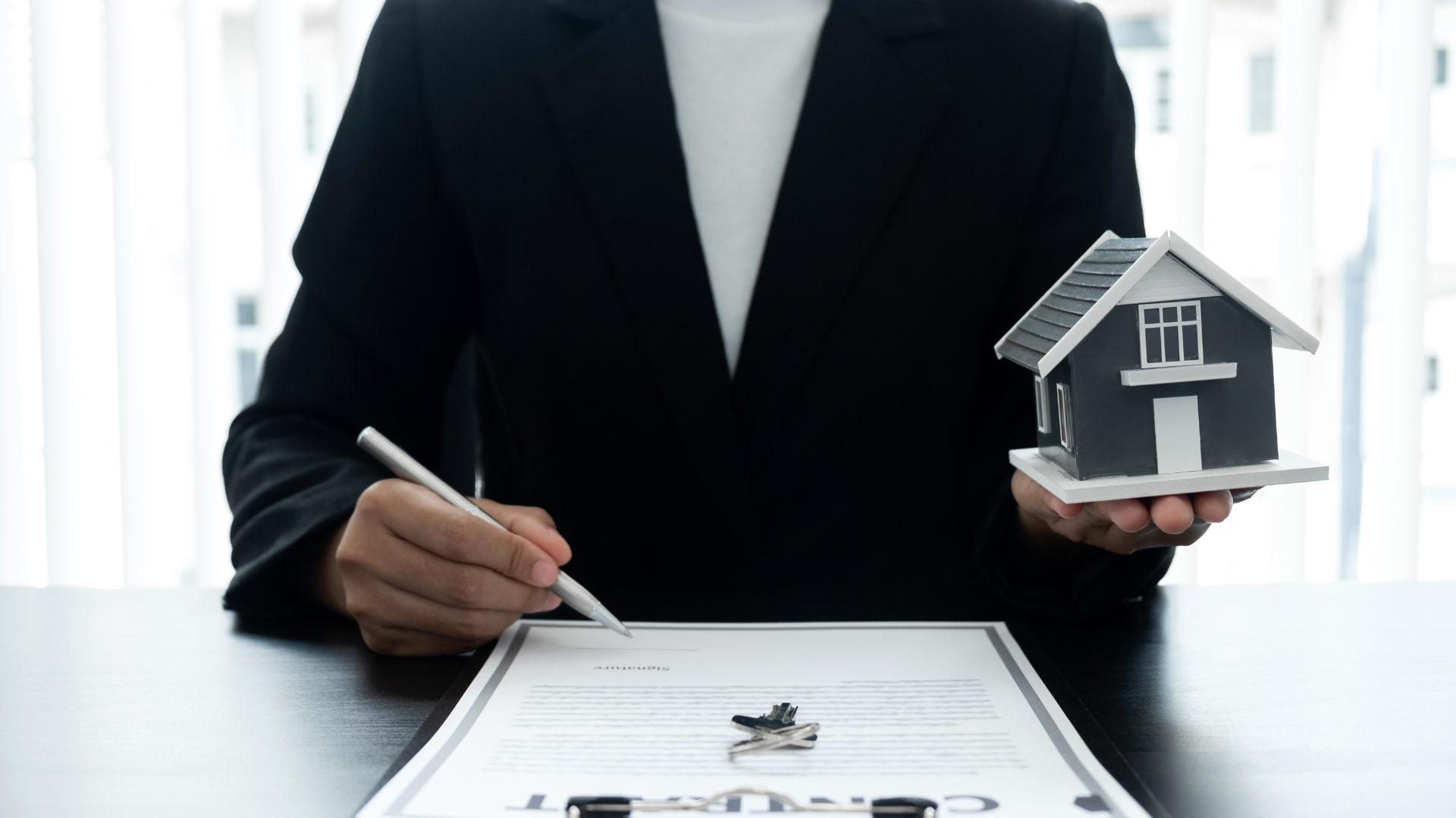 Une personne en costume signe un document, tenant une maison miniature, à un bureau.