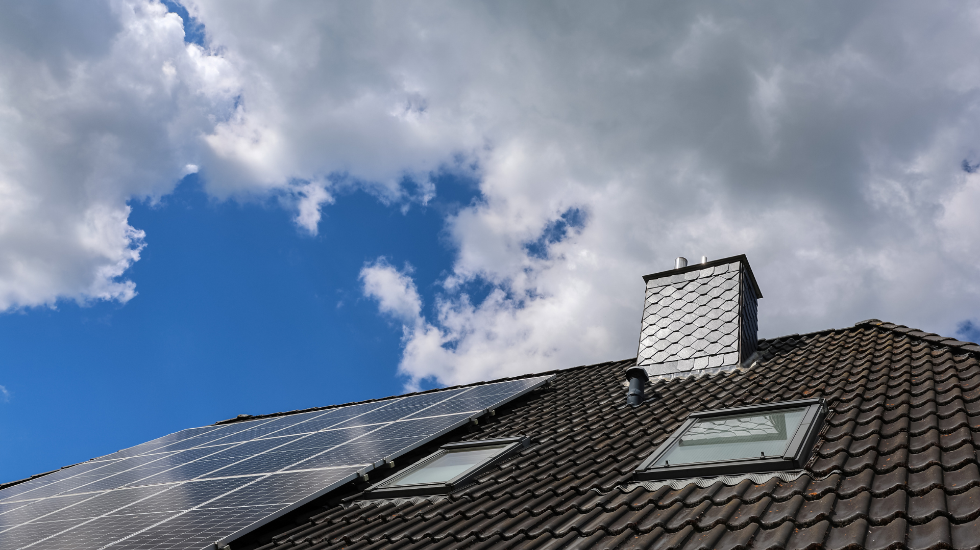 Panneaux solaires sur un toit sombre, avec cheminée et puits de lumière, sous un ciel bleu partiellement nuageux.