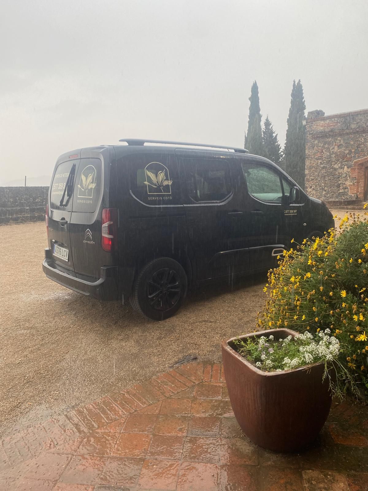 Furgoneta negra aparcada davant d'un edifici de pedra. La pluja cau sobre un pati de maó amb testos amb flors.