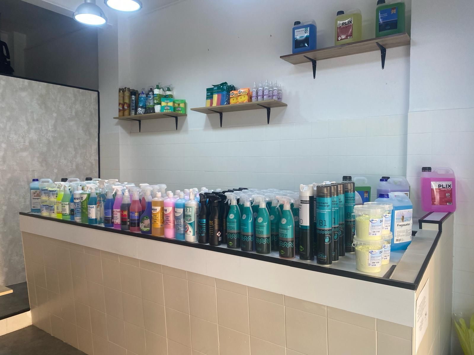 Un espai comercial amb prestatges plens de productes de neteja. Un taulell mostra ampolles de diversos colors.