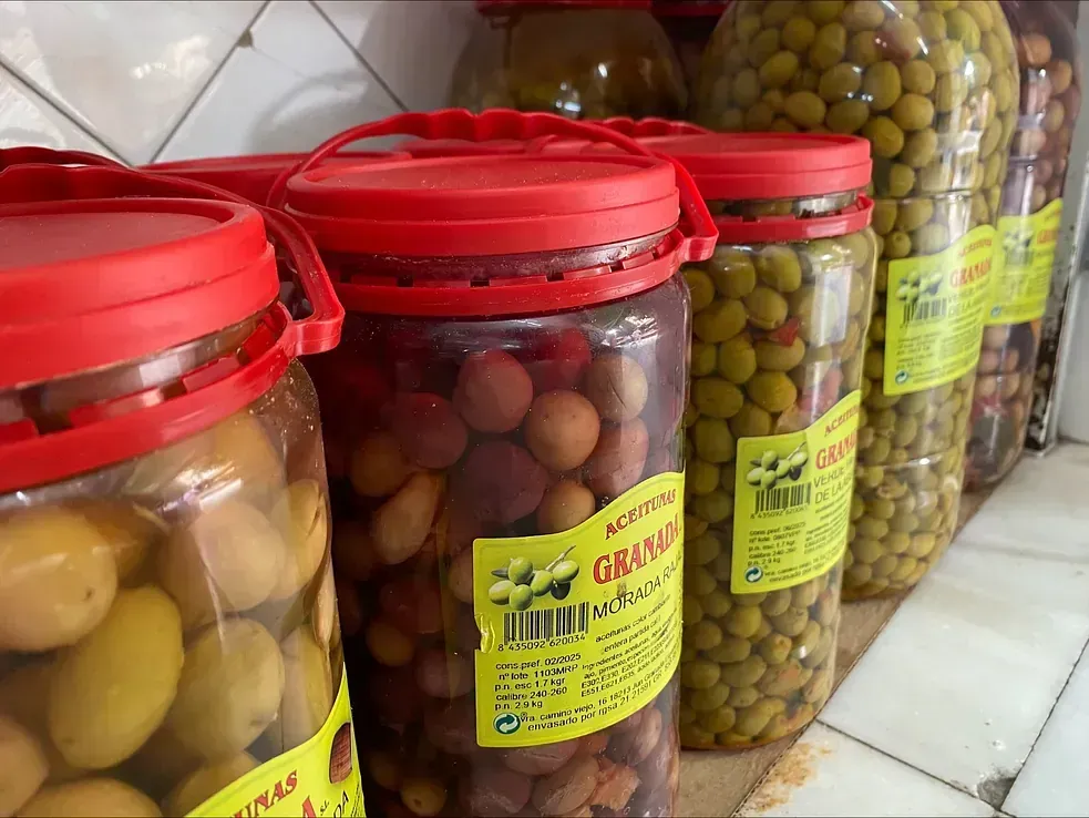 Tarros de aceitunas encurtidas con tapas rojas en un estante, de varios colores, algunos con etiquetas.