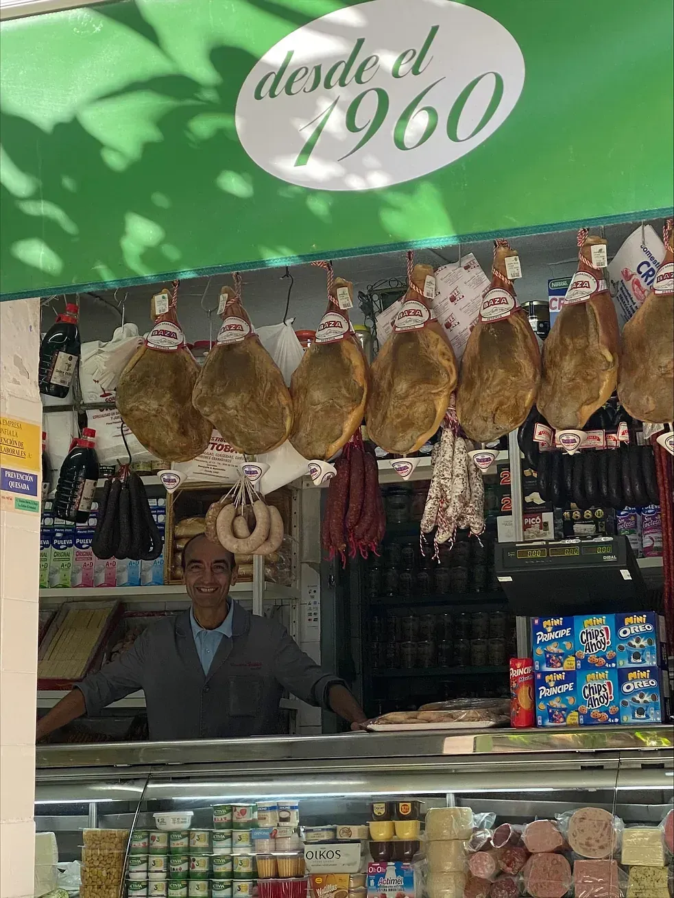 Hombre en un mostrador de charcutería con jamones, quesos y otros productos curados colgados. Letrero