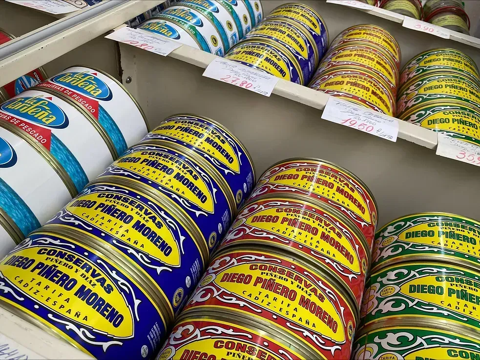 Comida enlatada exhibida en el estante de una tienda, con etiquetas de colores: azul, amarillo, rojo, verde.