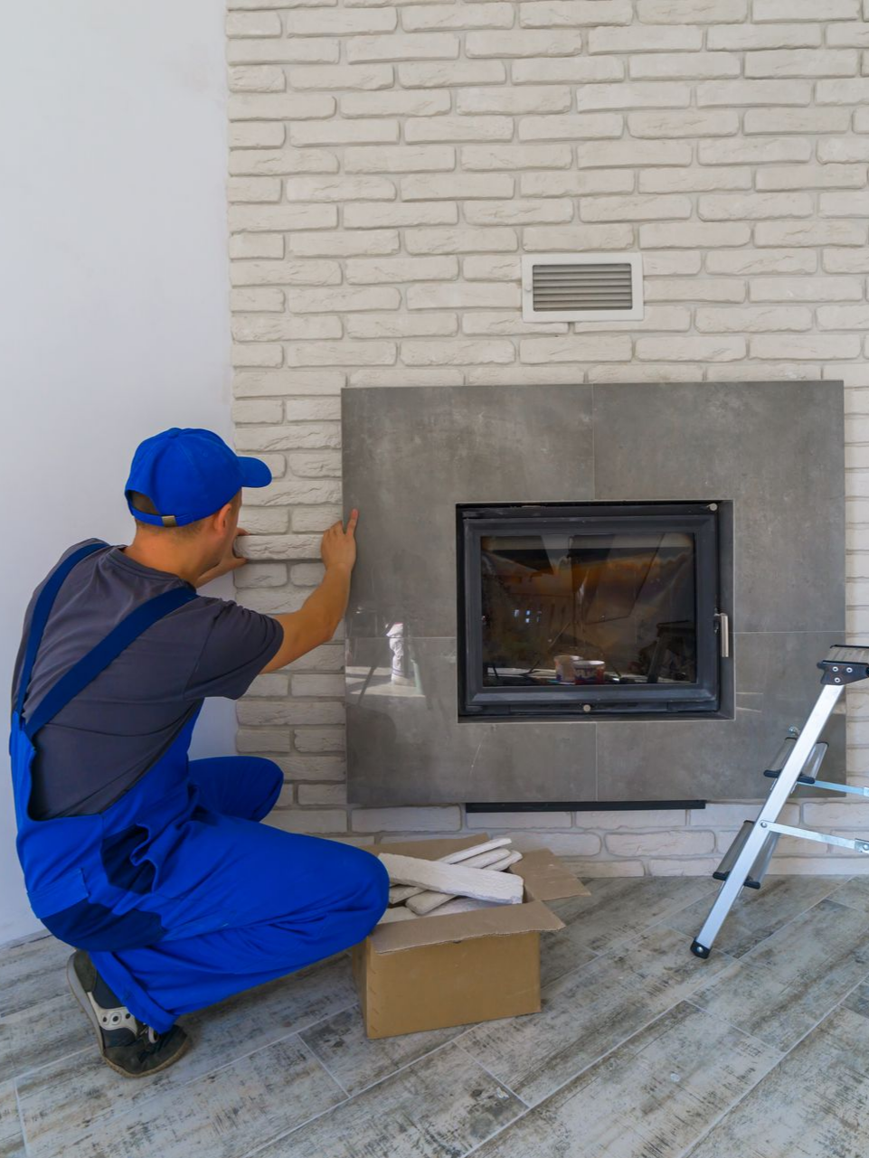 Personne en combinaison bleue posant du carrelage autour d'une cheminée sur un mur de briques.