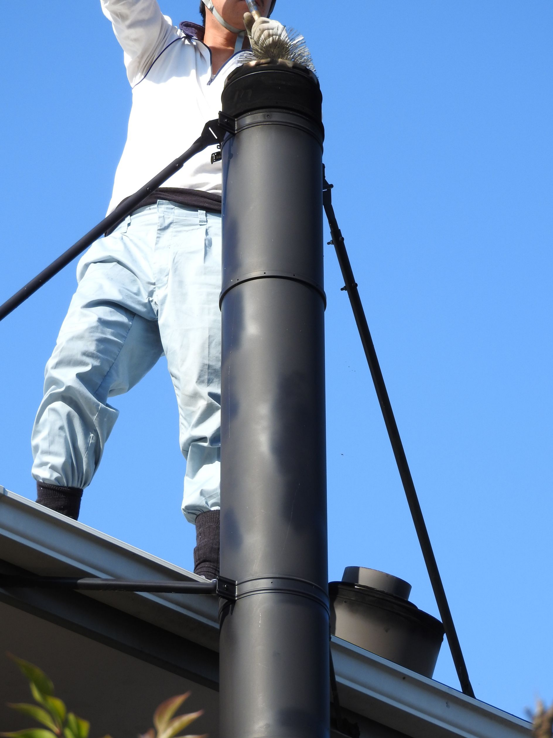 Une personne sur un toit nettoie une cheminée noircie avec une brosse, sur fond de ciel bleu.
